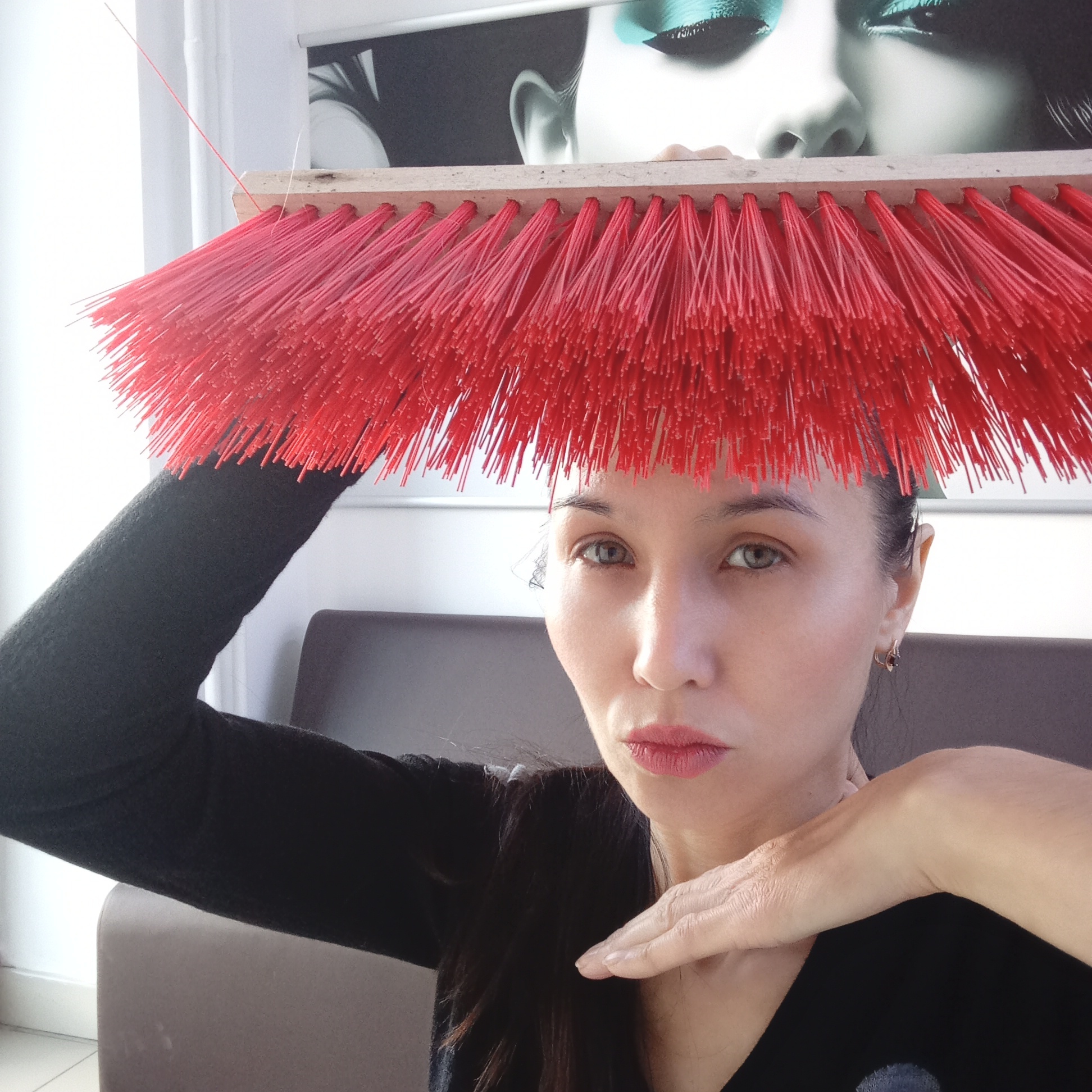 A woman with light skin, dark hair, and green eyes is posing indoors, holding a red-bristled broom above her head. She is wearing a black top. Her left arm is raised to support the broom while her right hand is positioned near her neck. The background includes a wall poster depicting a woman with dramatic eye makeup. Behind the woman is a tan cushioned object. The location is within a building, presumably in Izhevsk, Russia, although there are no specific environmental markers to definitively confirm this.