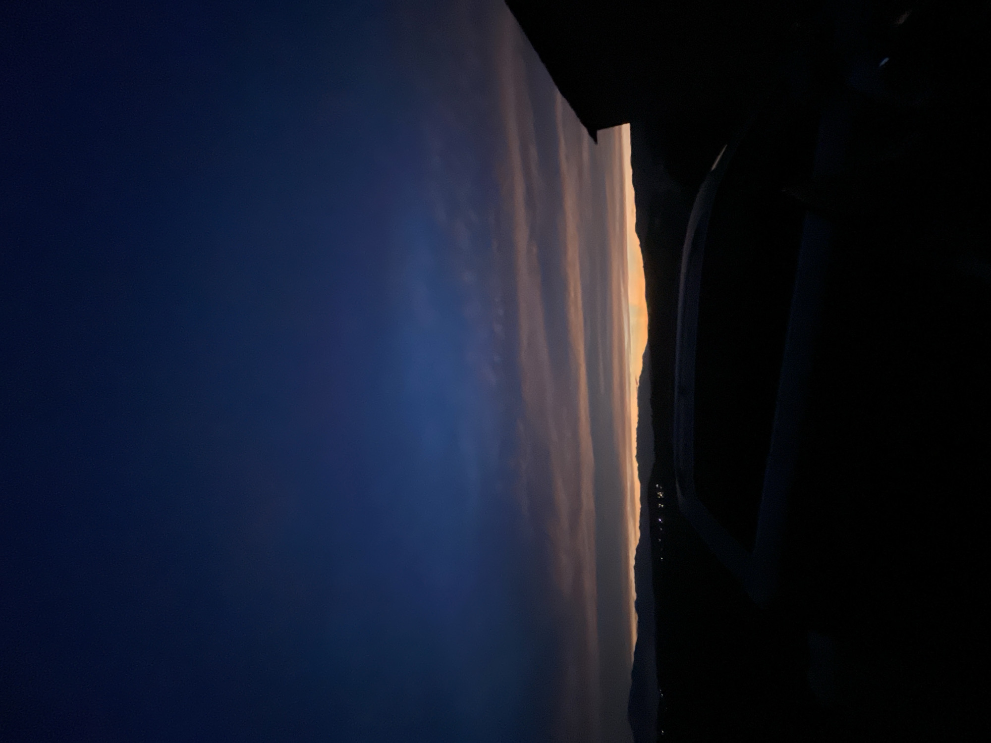 A vertical, low-light photograph captures a silhouette of a vehicle in the foreground against a dusky sky. The sky transitions from a deep, dark blue at the top to bands of soft pink and orange near the horizon, indicating either dawn or dusk. A faint line of distant mountains is visible along the horizon, with scattered lights suggesting a settlement. To the right, the dark silhouette of a building is partially visible. The overall scene suggests an outdoor setting in the city of Bogolin, Bulgaria.
