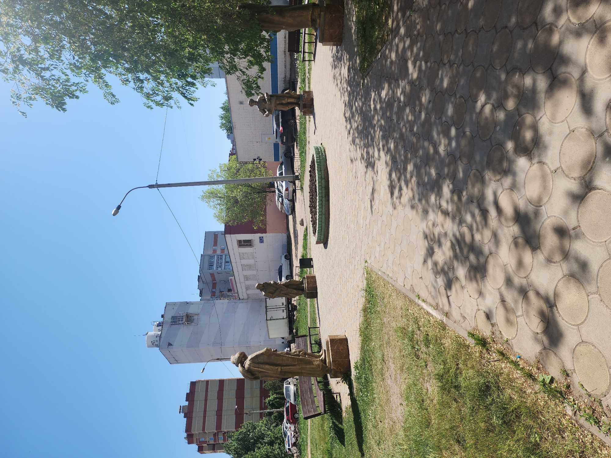 The image shows a paved path in Kazan, Russia, lined with statues of figures dressed in robes. The path consists of circular paving stones, and shadows from trees overlay its surface. Adjacent to the path is a strip of grass and several benches. In the background are multi-story buildings of various colors, a street lamp, and parked cars. The sky is clear and blue. The statues are bronze in color and appear to be of historical figures. A low flowerbed or planter is visible along the path.