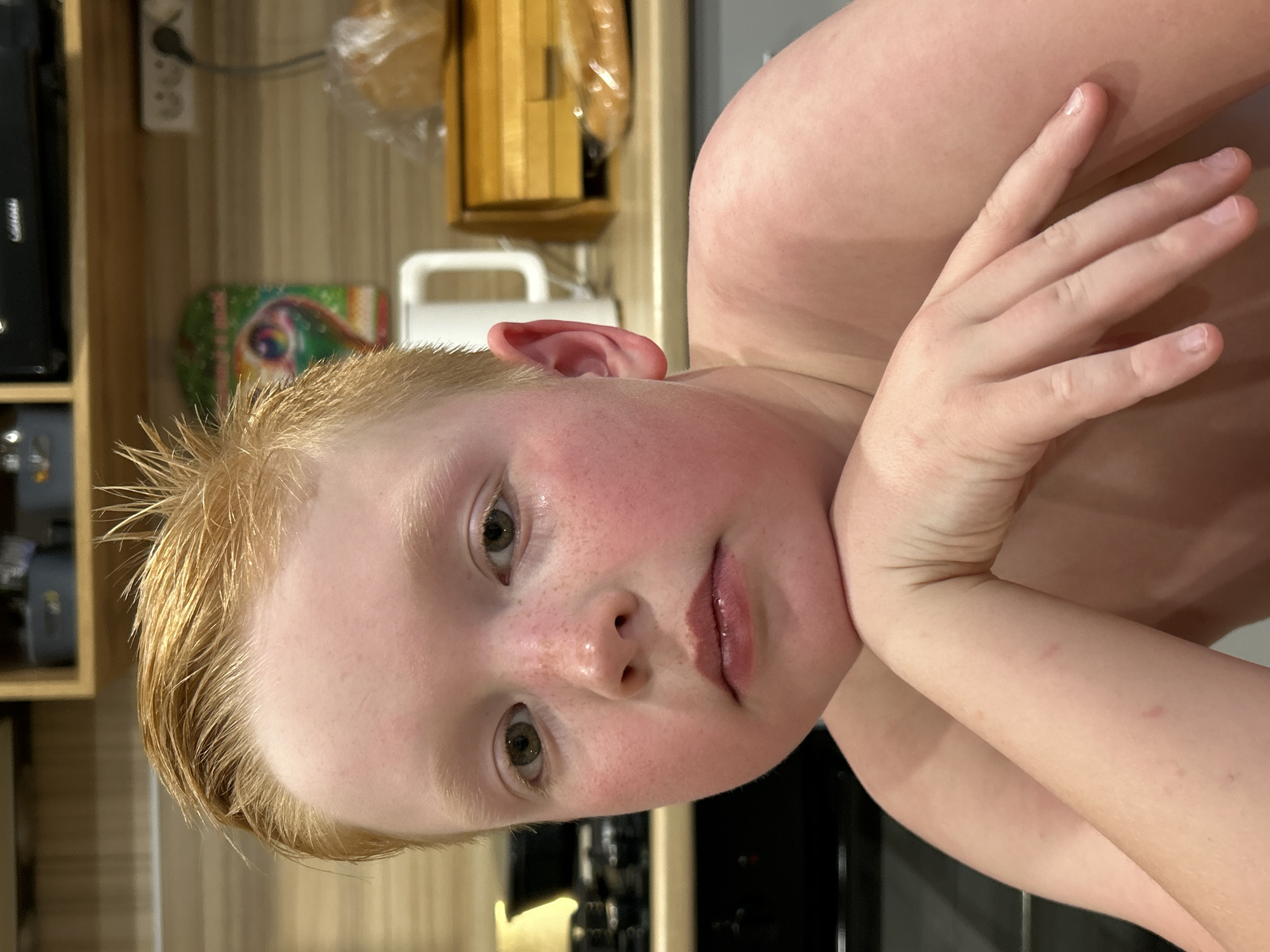 A fair-skinned, blond-haired child rests their chin on their hand, posing for a portrait. Freckles are visible on their face. The background reveals a kitchen setting with wooden cabinets and a few appliances. The location in the city of Уральское, Russia cannot be determined from the visual content.