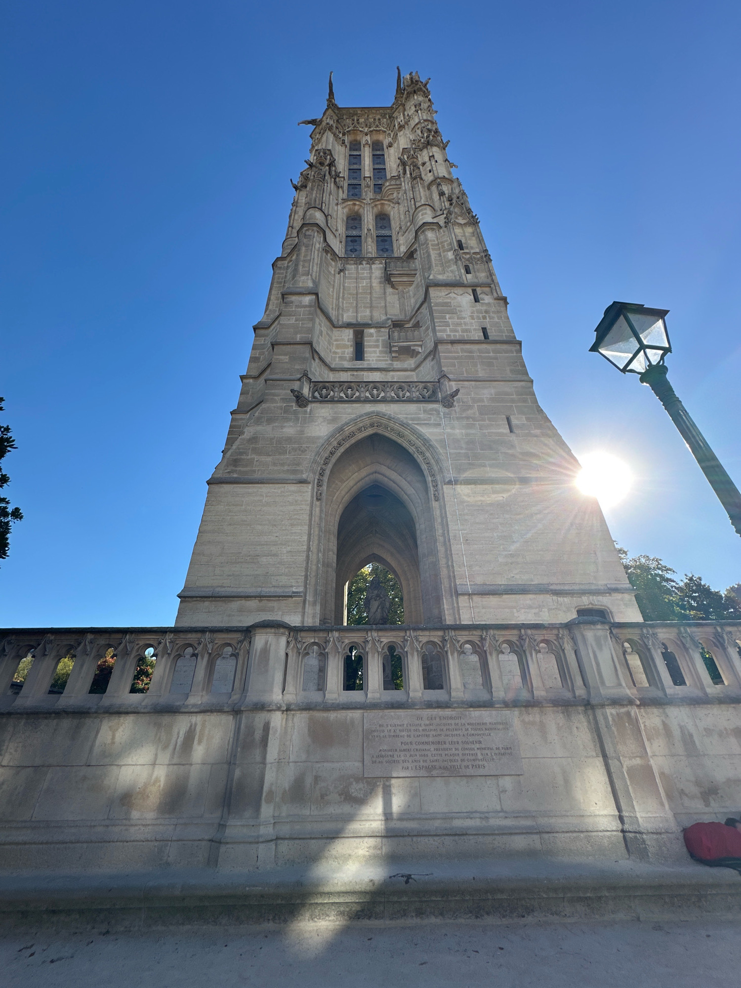The image shows the Tour Saint-Jacques in Paris, France, against a clear blue sky. The gothic tower is made of stone and is topped with spires and gargoyles. In the foreground, there is a stone balustrade with decorative carvings. A plaque is affixed to the lower part of the balustrade with French text. To the right of the tower, a streetlamp with an ornate base is visible, and the sun is shining brightly behind it, creating lens flare. Trees with green and red foliage are present to the left and right of the tower. The ground in the foreground appears to be paved, with shadows cast by the tower and balustrade. A red bag and a dark object, possibly a blanket, are on the ground near the lower right corner of the image. The perspective is from a low angle, looking upwards at the tower.