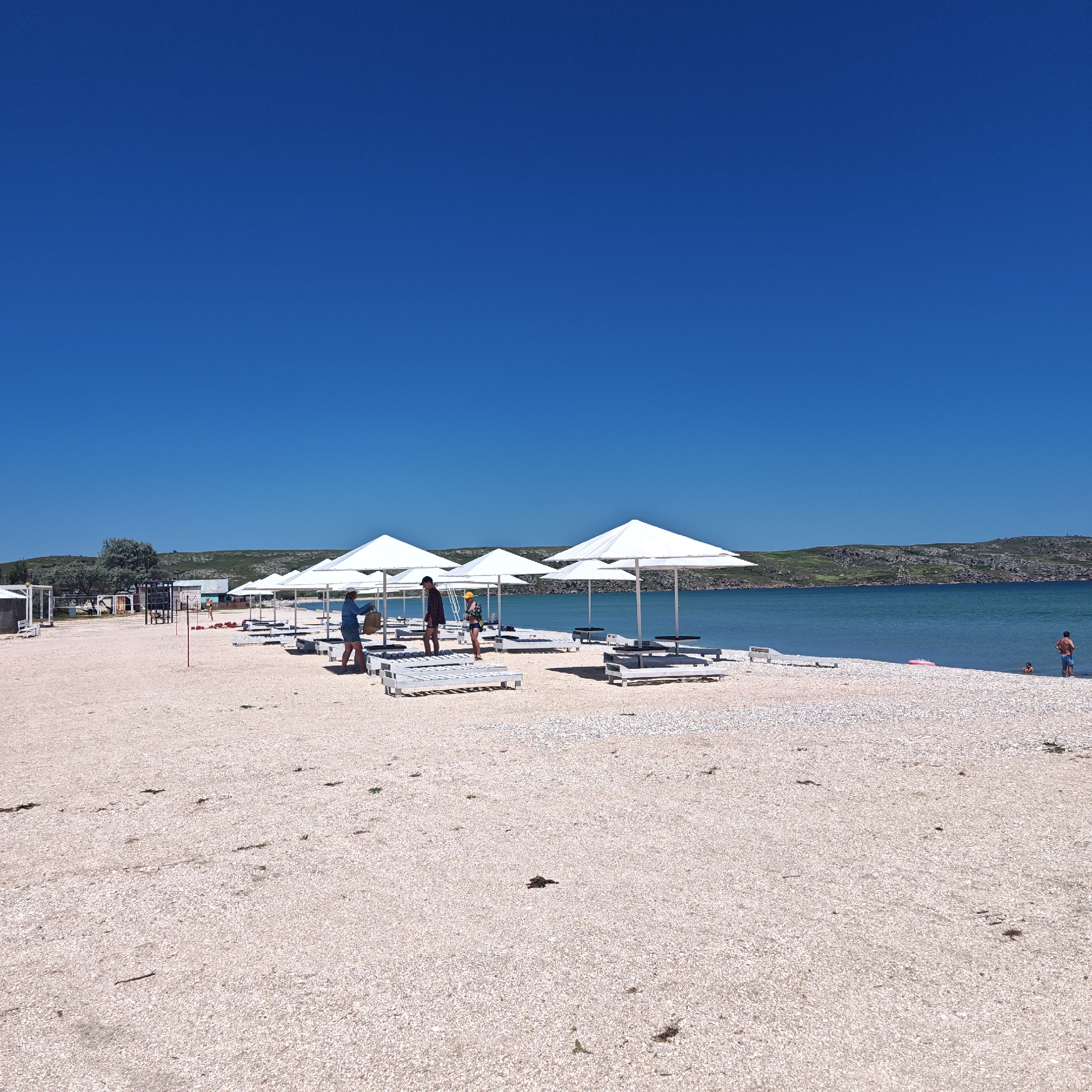 The image shows a beach in Unknown, Unknown, with a bright blue sky. The beach is covered in small white pebbles and shells. Several white sun umbrellas are arranged in a row, with white lounge chairs underneath. A few people are walking on the beach, and a couple of people are in the water. In the background, there are hills and a body of water. The overall impression is of a sunny day at a resort or vacation destination.