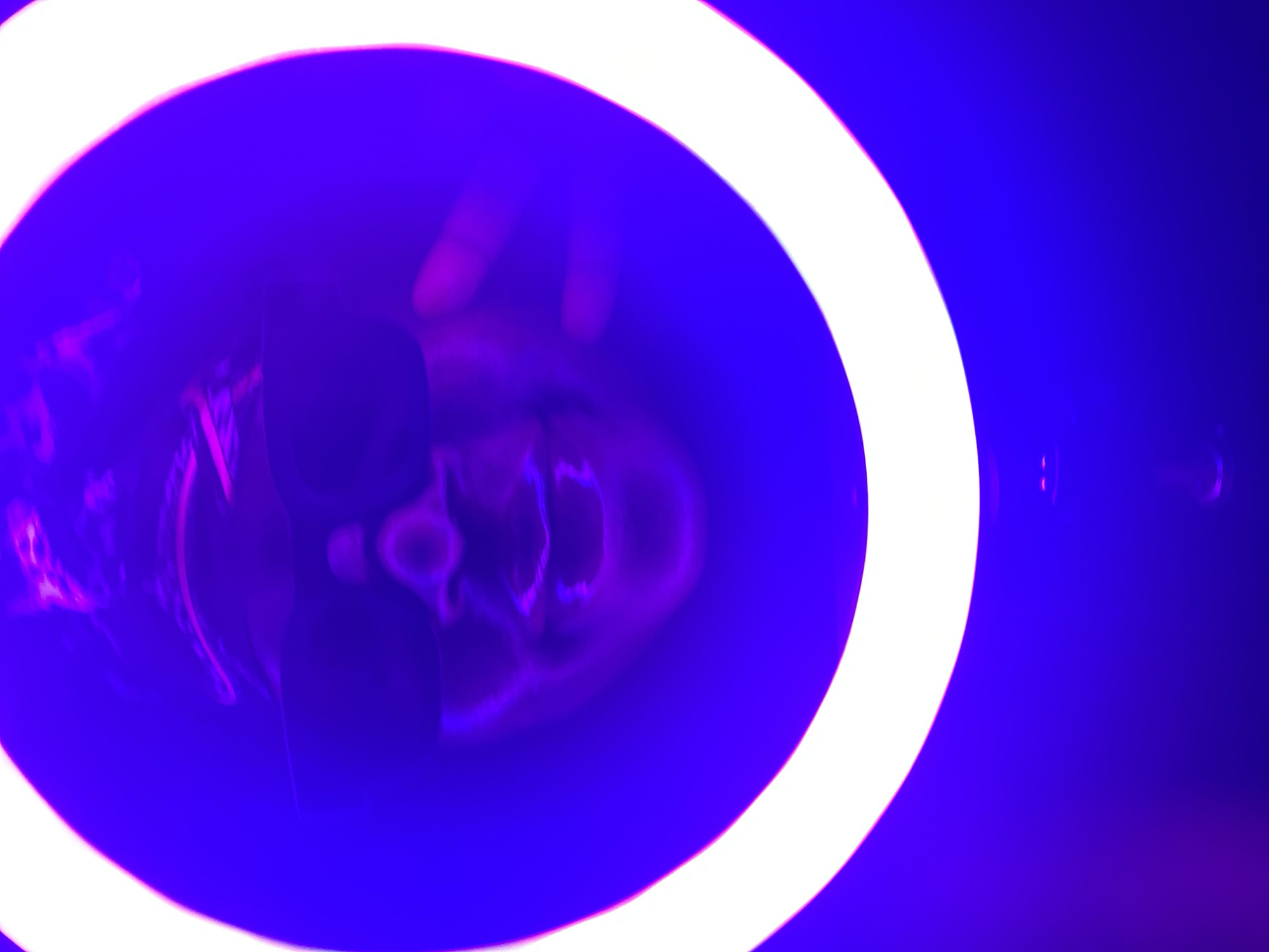 The image captures a surreal and abstract scene dominated by vibrant purple and blue hues, with a prominent white ring light framing the center. The primary subject appears to be a distorted human face or head, visible within the circle of the bright white light. The facial features are blurred and stretched, suggesting a reflection or a view through a distorting medium. On the left side of the frame, blurry outlines resembling fingers or a hand are visible, seemingly interacting with the surface or object causing the reflection.
The setting is characterized by its intense artificial lighting, creating a deep, almost glowing blue-violet background. The circular white light source casts a strong illumination, contrasting sharply with the darker, colorful environment. There are no clear cues regarding the specific time of day or weather, but the scene is distinctly lit by artificial sources, suggesting an indoor location or nighttime. The overall mood is mysterious and artistic due to the distortion and saturated colors. No visible text is present in the image. This scene takes place in Maigizo, Nigeria.