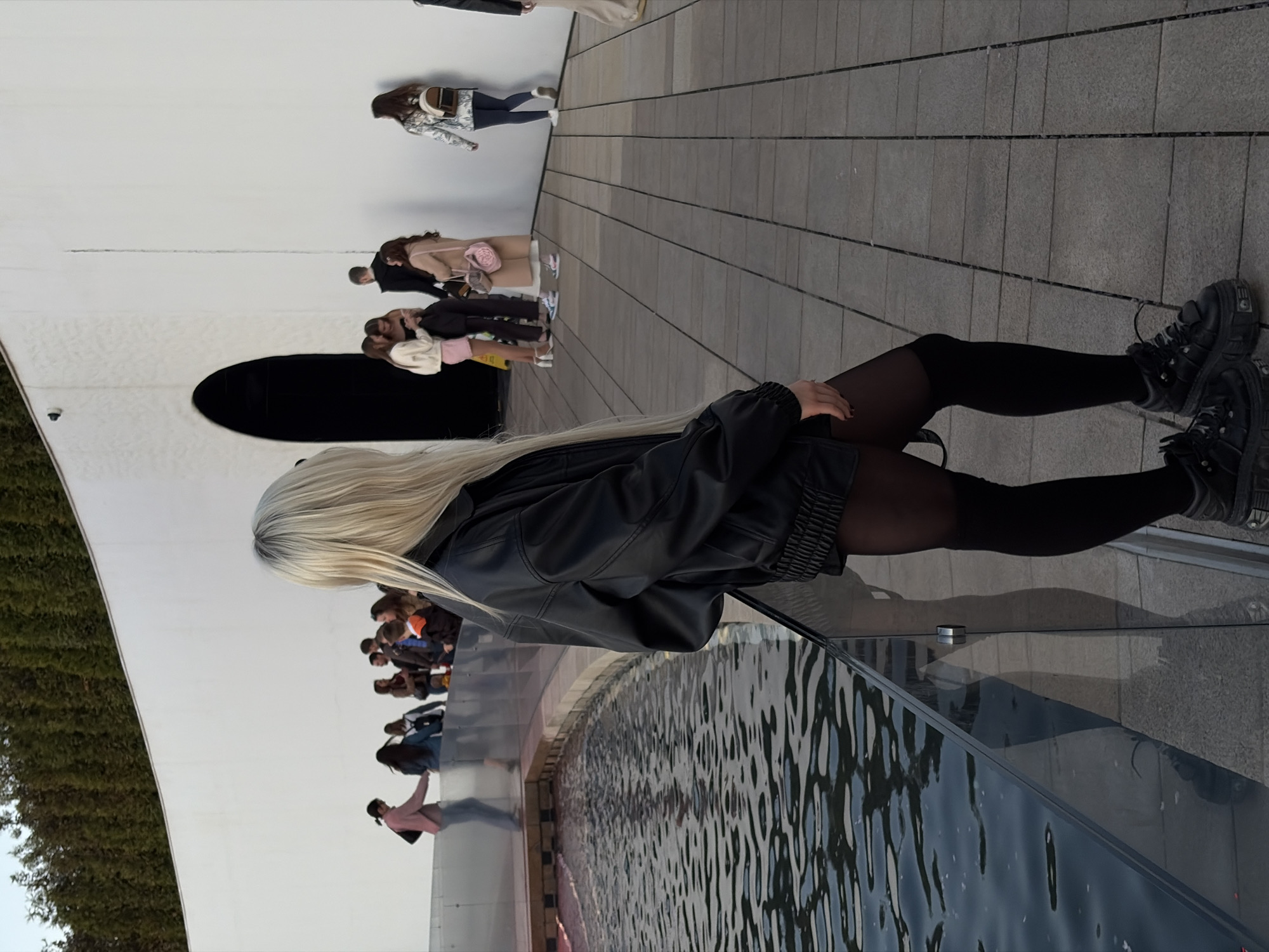 The image shows a woman with blonde hair and a black leather jacket standing near a water feature or fountain. She is wearing black tights and black platform shoes. The background reveals an outdoor location with a large white curved wall. Other people are visible near the wall, suggesting a public space or attraction. The scene is near the Galitsky Park in the city of Krasnodar, Russia.