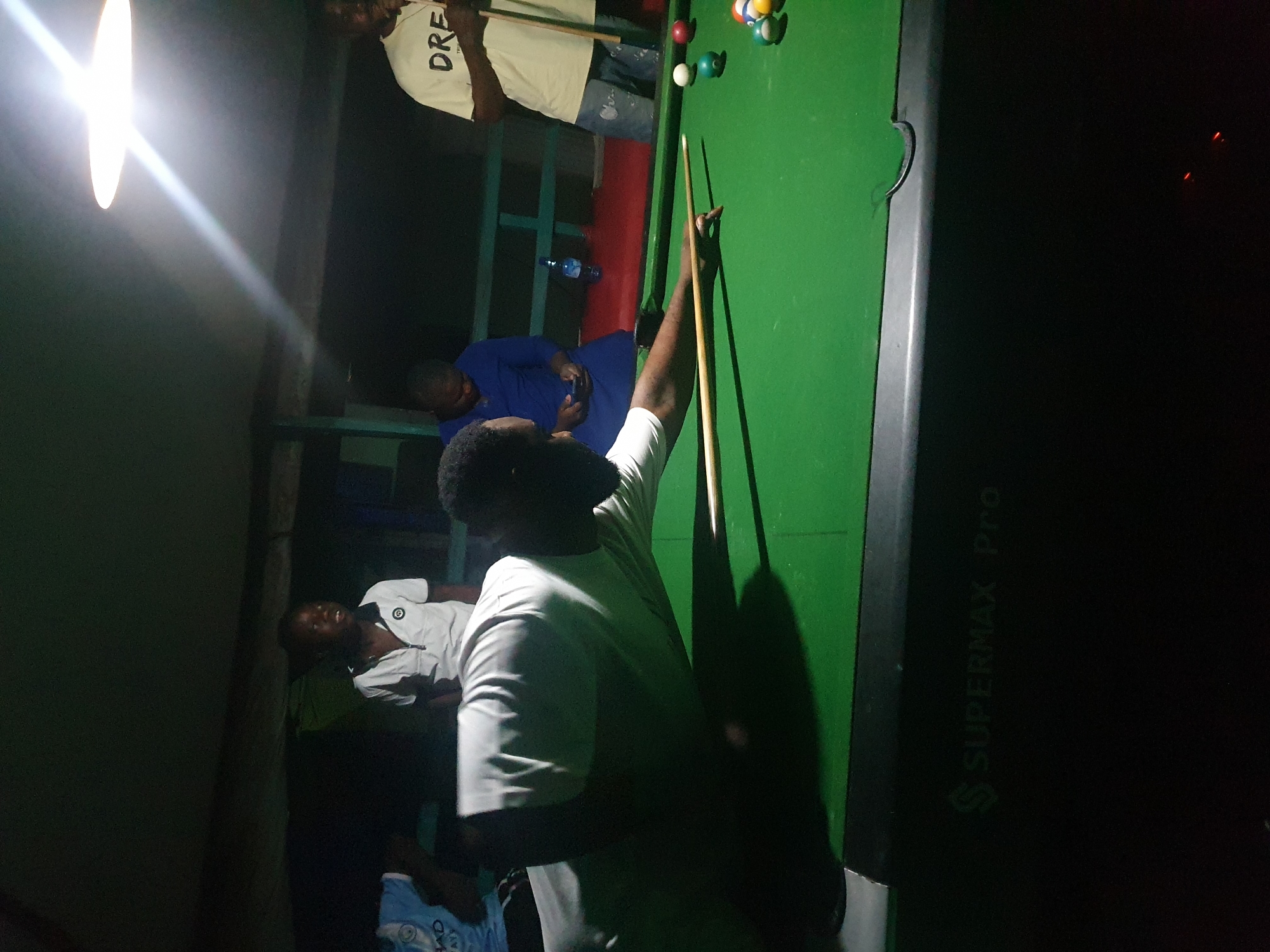 In Lokoja, Nigeria, a group of individuals are gathered around a green pool table under artificial light. A Black man, the primary subject, is in the foreground, leaning over the table with a pool cue in his right hand, preparing to strike a ball. He is wearing a white t-shirt. To his right, on the pool table, are several colored pool balls, including a white cue ball, a green ball, and a cluster of other balls. In the background, a young Black boy in a white collared shirt is visible, looking towards the viewer. Next to him, a Black woman in a blue dress is looking down at her phone. Further to the right, another Black man in a white t-shirt with the word "DRE" printed on it stands holding a pool cue. The environment appears to be an indoor or semi-outdoor space with a ceiling light fixture illuminating the scene. The pool table itself has the brand name "SUPERMAX Pro" embossed on its side. The overall atmosphere suggests a casual gathering focused on playing pool.