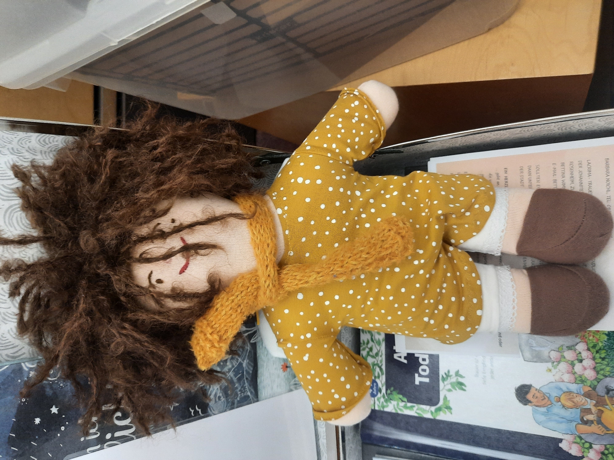 A stuffed doll with frizzy brown hair and a yellow polka-dot dress sits upright, its legs propped on what appears to be a book. The doll is wearing a knitted yellow scarf around its neck, white socks with lace trim, and brown shoe-like coverings on its feet. The background is a mix of blurred objects, including a white plastic bin on the right and what seems to be book covers with text and illustrations on the left. One of the book illustrations depicts a man and a child tending to flowers, with German text visible, including "DER STADT" and "ROSENHEIM". The context of the image, with the German text and location reference, suggests it was taken in Rosenheim, Germany.
