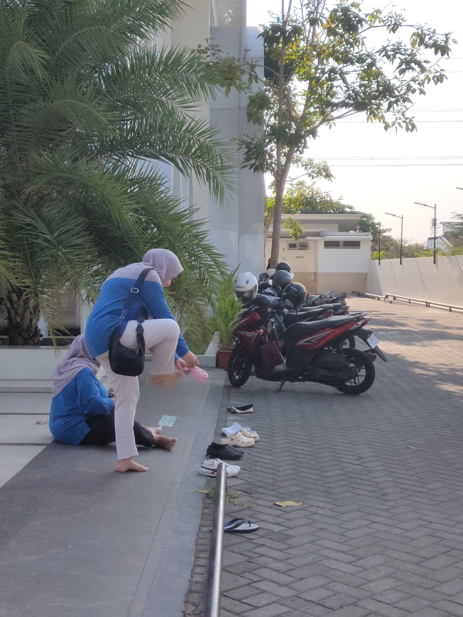 In the city of Ngasem, Indonesia, two individuals, both wearing hijabs and modest attire, are present on an outdoor paved area. One individual is seated on a low ledge, facing away from the camera, and is wearing a blue top and dark pants. The other individual is standing, leaning forward, and appears to be tying or adjusting their shoes, wearing a blue and light-colored top with white pants and a black crossbody bag.
Several motorcycles are parked in a line on the right side of the image. Scattered on the paved ground in front of the motorcycles and near the seated individual are various pairs of footwear, including sandals, flip-flops, and closed-toe shoes.
The environment includes a building with white walls and windows in the background, alongside a tall, slender tree. Lush green palm fronds from a large palm tree are visible on the left side of the frame, partially obscuring the building. The lighting suggests it is daytime, with sunlight casting shadows. The ground is paved with interlocking bricks.