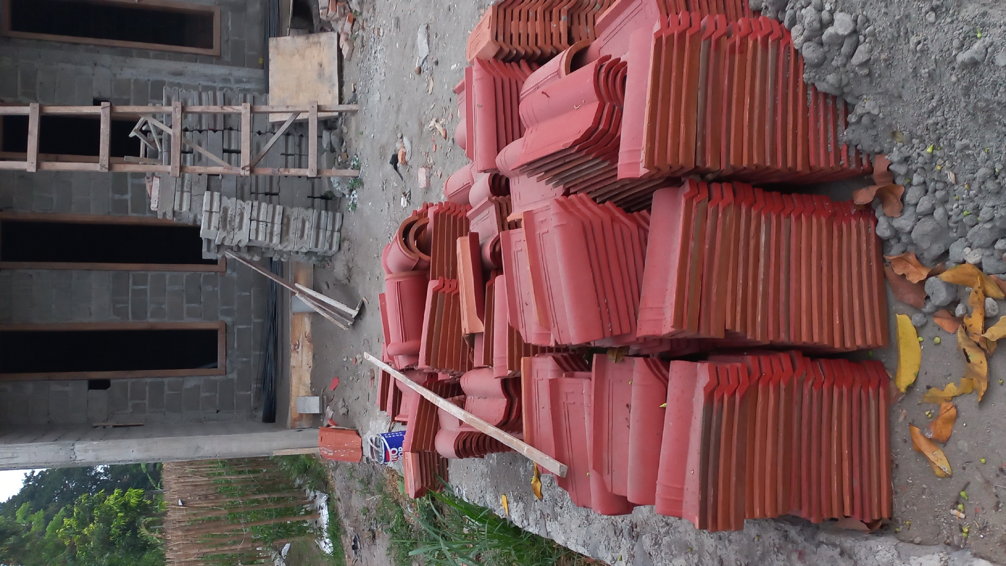 The image depicts a construction site in Pare, Indonesia, with a partially built house in the background. The house is constructed with grey concrete blocks and has two dark, rectangular window openings on the visible facade. To the right of the house, a wooden ladder leans against a stack of grey bricks. In the foreground, a large pile of red roof tiles is stacked on the ground. A wooden plank is resting on top of some of the roof tiles, and a metal can, possibly paint or primer, is also visible near the tiles. The ground is a mix of dirt, gravel, and scattered dry leaves. A bamboo fence runs along the left side of the image, bordering the construction area. The overall scene suggests ongoing construction work in an outdoor setting.