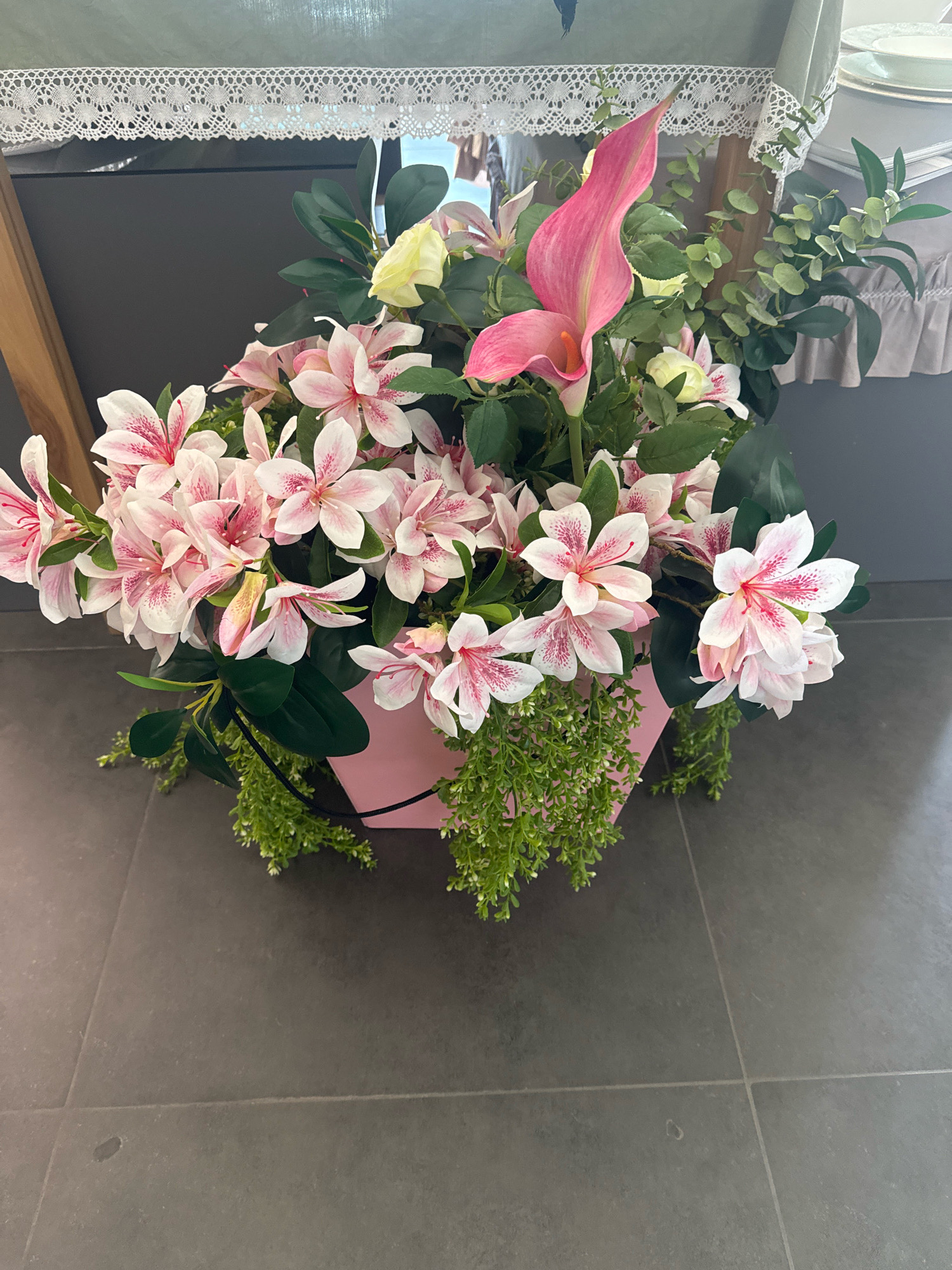 A floral arrangement in a pink hexagonal pot sits on a dark gray tiled floor. The arrangement features numerous light pink flowers with darker pink speckles, one prominent bright pink calla lily, and various green foliage. The arrangement is situated in front of a dark gray wall, with a wooden table leg visible on the left. In the background, a tablecloth with white lace trim is draped over a table, and other tableware is partially visible. The scene appears to be indoors, possibly in a home or event space. The image does not contain any information that would allow for the identification of its location in Novosibirsk, Russia.