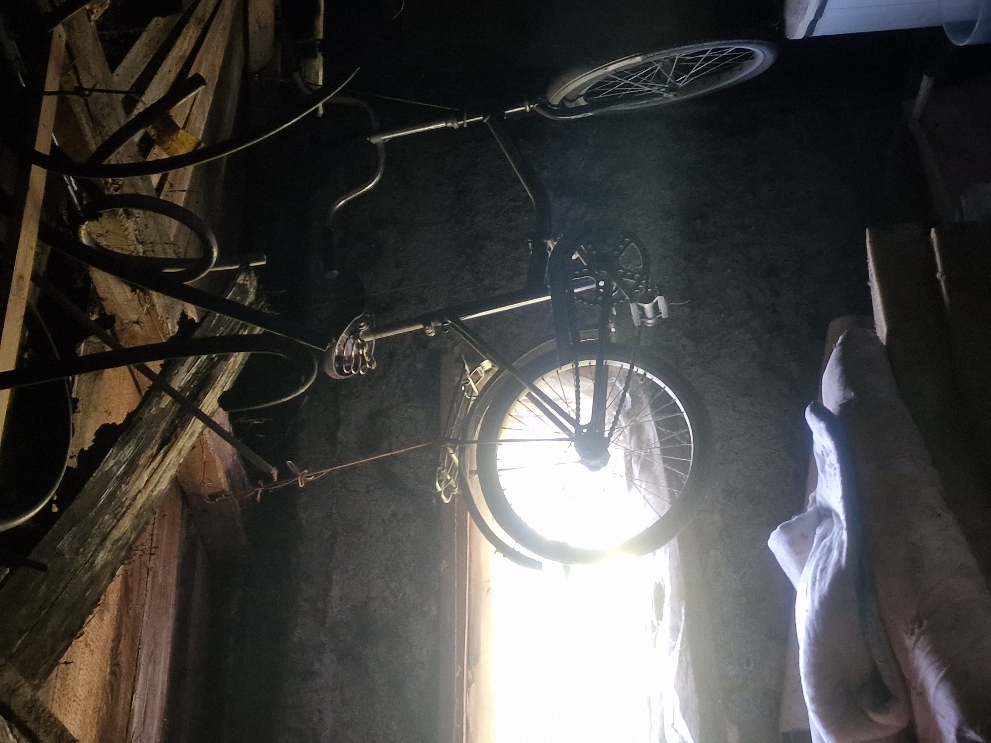 An old, black bicycle is suspended from the ceiling of a dimly lit, rustic interior, possibly a shed or garage, in Makinsk, Kazakhstan. The bicycle is hung by a rope from a wooden beam. Light streams in from a window on the left, illuminating the rear wheel of the bicycle and a white, blanket-like object resting on a surface below. The walls appear to be made of concrete or rough plaster. The overall scene suggests a storage area for unused items.
