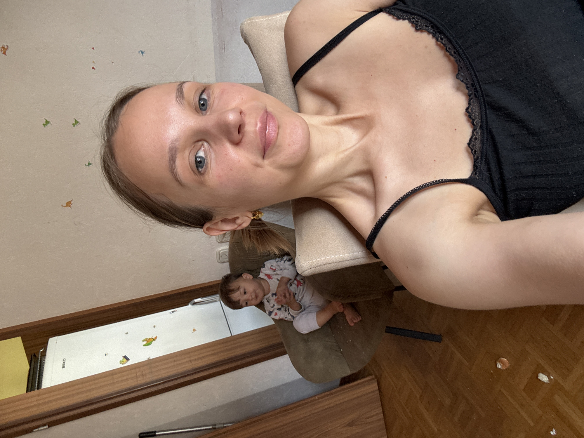 A young woman with blonde hair pulled back in a ponytail is seated in the foreground, looking at the camera. She is wearing a black camisole with lace trim. Her left arm is extended towards the camera, and her right arm is bent, resting on a beige chair. Behind her, to the left, a young child is seated in a brown armchair, facing to the left and looking away from the camera. The child is wearing a long-sleeved pajama top with a pattern of animals and white pants. The background includes a white refrigerator with colorful stickers on it, and wooden doors. The floor is made of light brown parquet. The overall scene appears to be indoors, possibly in a residential setting, in Überherrn, Germany.
