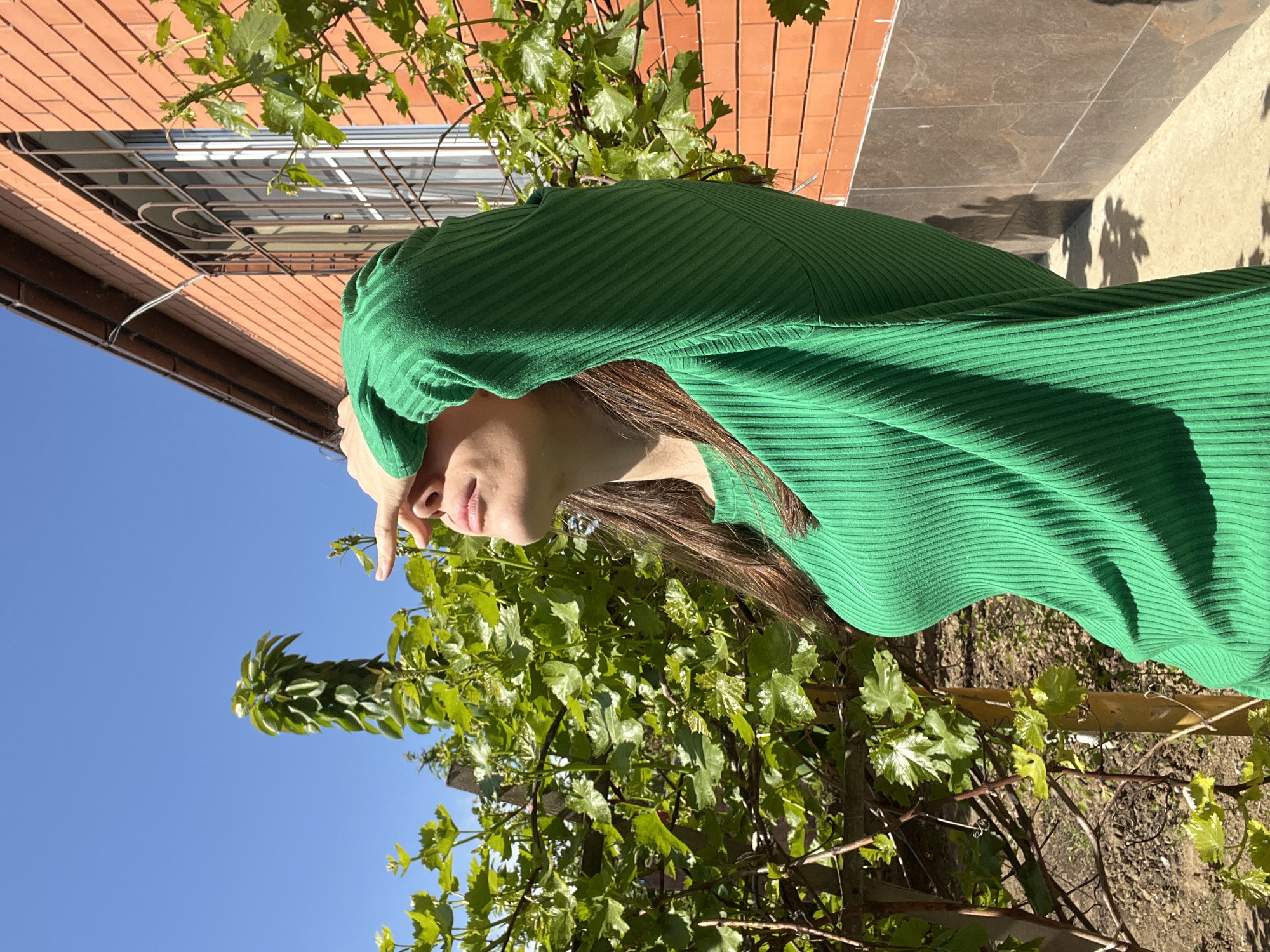 A woman in a green, ribbed, long-sleeved garment is the central subject, standing outdoors in what appears to be Kaspiysk, Russia. Her right hand is raised to her forehead. The backdrop includes a brick building, foliage including vines and trees, and a clear blue sky in the background. A window with a metal grill is visible on the brick building. The ground appears to be paved, with some visible shadowing.