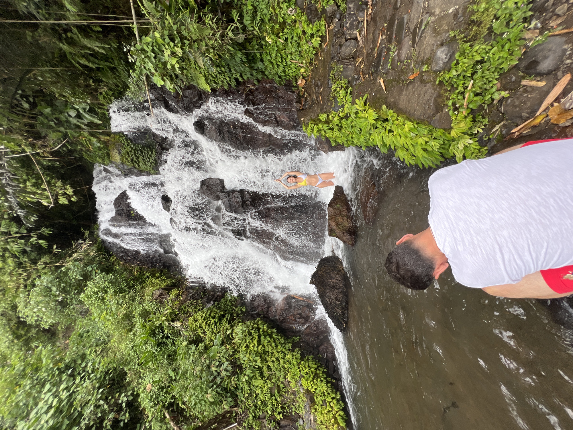 In Sukasada, Indonesia, a waterfall cascades down dark, moss-covered rocks, surrounded by lush green foliage. A person in a white t-shirt and red shorts sits in the foreground, facing away from the camera, looking towards the water. In the middle ground, a woman stands in the shallow water at the base of the waterfall, wearing a yellow and white bikini. She has her hands raised above her head, palms together. The water is clear and reflects the surrounding greenery. The scene is natural and serene.