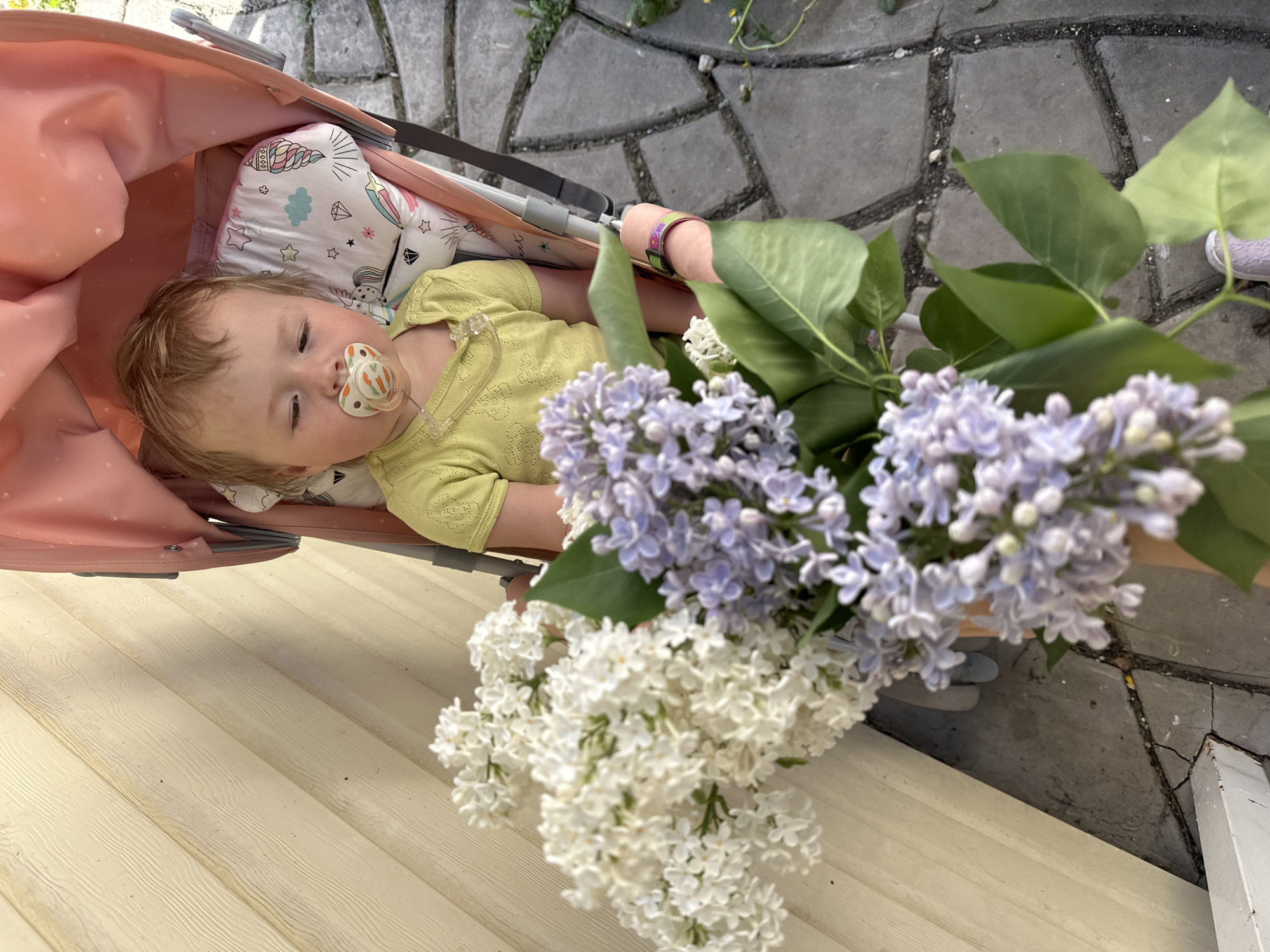 A baby lies in a stroller with a pacifier in its mouth. The baby is wearing a light green outfit and is resting on a white pillow with colorful designs. Lilac flowers are placed next to the baby in the stroller. The stroller sits on a light-colored wooden surface, which appears to be a porch or deck. The background features a stone pathway or patio. The city and location are not identifiable based on the image.