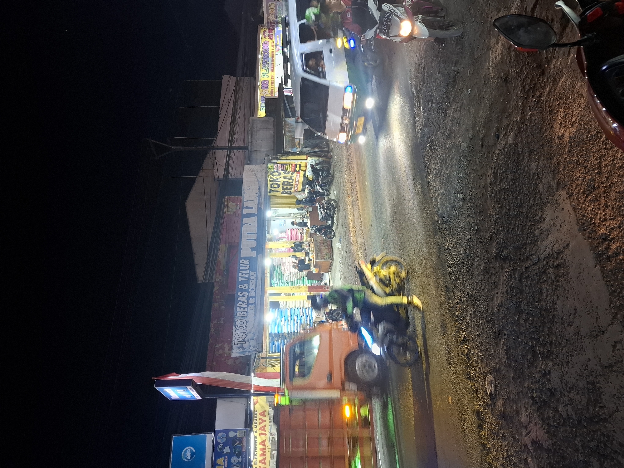 This is a nighttime, eye-level, medium shot of a street scene in Karangtengah, Indonesia. The road appears to be paved with asphalt and gravel, with visible potholes and uneven surfaces. Several vehicles are present, including a bright orange truck with its hazard lights on, a white van with green trim driving on the right side of the road, and multiple motorcycles.
On the left side of the street, a store with a blue sign that says "ELEKTRONIK & MEUBEL" and "KAYAMA JAYA" is visible, along with a red and white Indonesian flag. Next to it is another store with a blue sign that reads "TOKO BERAS & TELUR PUTRA LANGGENG". Inside this store, stacks of rice sacks are visible. Further down the street, a store with a yellow sign that says "BINTANG PLAYGROUND" is illuminated.
Several people are depicted in the scene. A person is riding a motorcycle with a yellow body and black accents in the foreground, their face obscured by a helmet. Another person is riding a silver and pink motorcycle on the right side of the road. Inside the "TOKO BERAS & TELUR" store, two figures can be seen. The lighting suggests artificial streetlights and vehicle headlights illuminating the scene. The overall atmosphere is that of a busy commercial street at night.