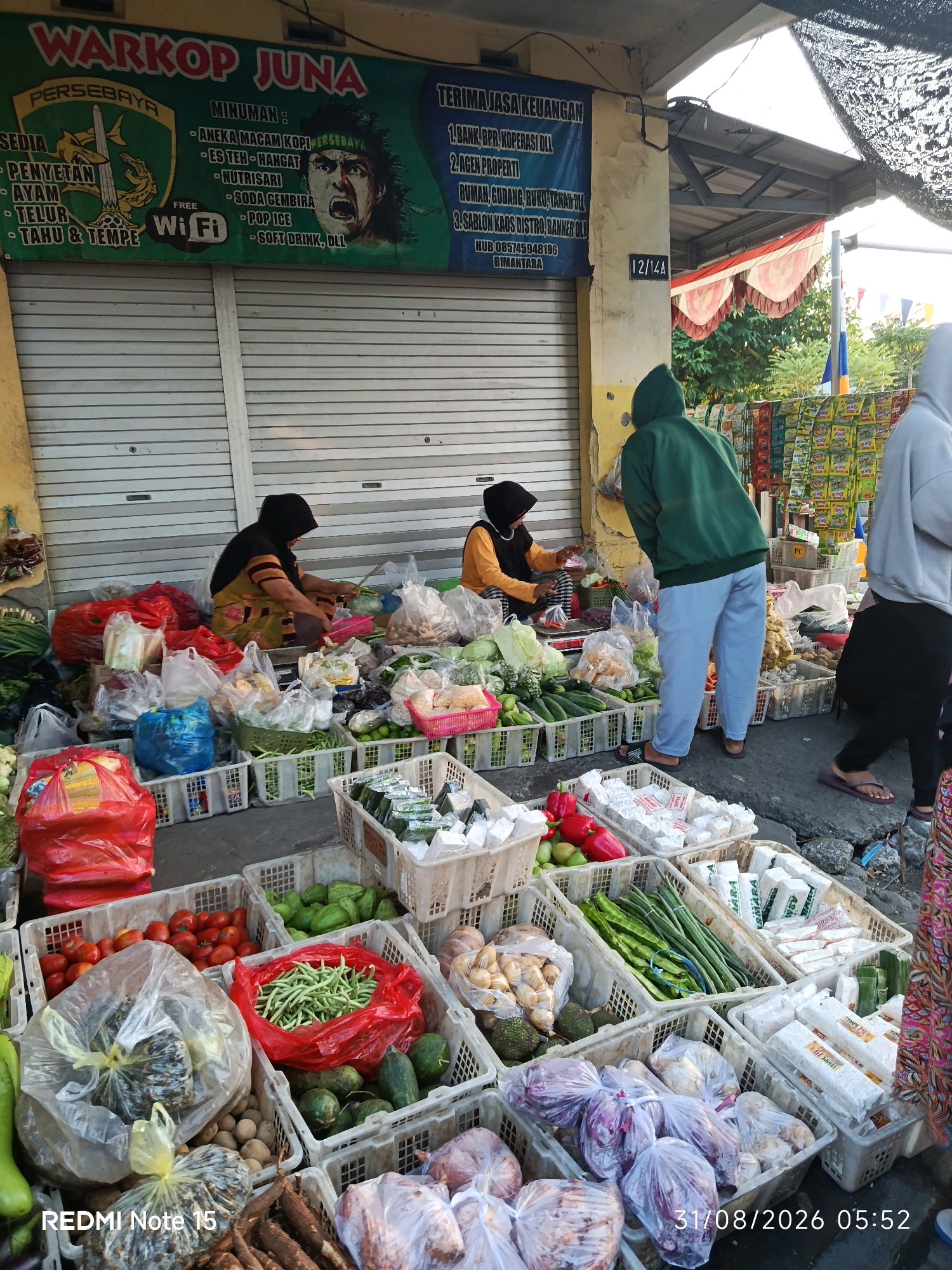The image captures an outdoor market stall, likely in Keboansikep, Indonesia, as indicated by the text "WARKOP JUNA" and an emblem possibly related to a local sports team. The stall displays a variety of fresh produce and packaged goods. In the foreground, several crates are filled with vegetables such as tomatoes, cucumbers, long beans, and onions. Packaged tempeh, a traditional Indonesian soy product, is also visible in white wrapping. Behind the produce, two women are attending the stall. One, wearing a headscarf and traditional clothing, is seated and appears to be arranging vegetables. Another woman, wearing a yellow top and headscarf, is also seated and engaged in activity at the stall. Two other individuals, seen from behind, are walking away from the stall, one wearing a green hooded sweatshirt and blue pants, and the other a grey hooded sweatshirt and dark clothing. The market stall is set up against a building with closed metal shutters, suggesting it might be early morning or late afternoon. Overhead, a black mesh awning provides shade. The overall scene depicts a typical street market environment with vendors and customers interacting.