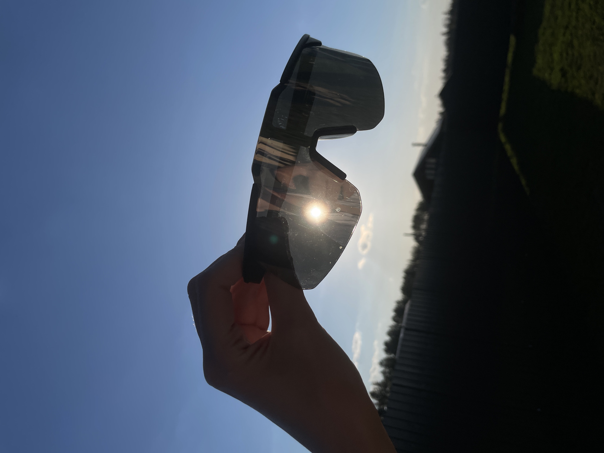 A person's hand holds a pair of dark-rimmed sunglasses up to the sun. The sun's light is visible through the sunglasses. The background consists of a bright blue sky on the left and a blurred, dark area of trees, grass, and possibly a building on the right. Based on the location of Подшивалово, Russia, it is likely a rural or suburban scene.