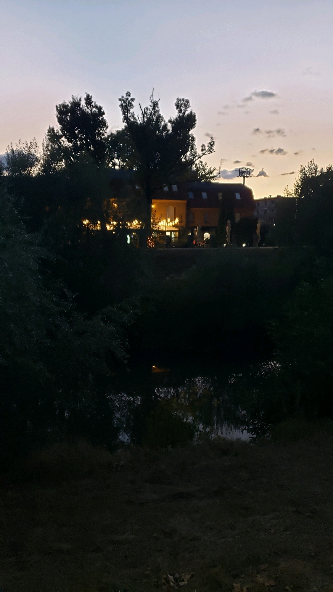 The image depicts an exterior view of a building at dusk, likely a restaurant or cafe, located in the city of Prešov, Slovakia. The building is illuminated from within, with warm yellow lights visible through windows and along an outdoor seating area. Several trees in the foreground and midground are silhouetted against the fading sky, obscuring parts of the building and its surroundings. A body of water, possibly a river or pond, is visible in the lower portion of the image, reflecting the dim light. The sky is a gradient of pale purple and orange, indicating the transition from sunset to twilight, with a few scattered clouds. The overall atmosphere is tranquil and serene, suggesting the end of the day.