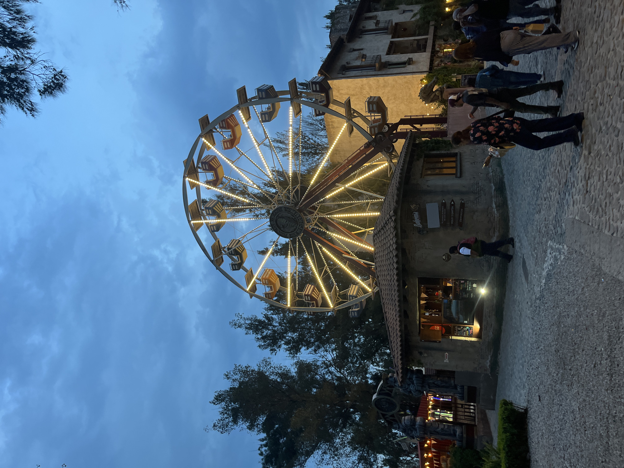The image depicts a Ferris wheel in Domingo Arenas, Mexico. The wheel is lit with string lights and has empty passenger cars. The sky is a dark blue color and partly cloudy. In the background, there is a building with lights on inside. There are several people standing and walking near the building. Trees are partially visible on both sides of the image. The ground is paved with stone. The image is oriented vertically.