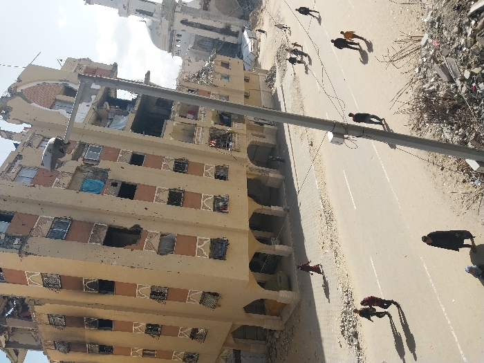 The image displays a street scene in Khan Yunis, Palestinian Territory, under bright daylight. A prominent, multi-story building, appearing to be at least seven floors high, occupies the left side of the frame. This building exhibits extensive structural damage, including collapsed sections of the roof and upper floors, missing exterior wall sections, exposed interiors, numerous shattered or absent windows, and black scorch marks around some window openings. Blue fabric hangs from an opening on a middle floor. To the right of this building, a mosque with a dome and a partially damaged minaret is visible, also showing signs of structural compromise.
A paved asphalt road, featuring dashed white lane markings, runs through the mid-ground. Debris, including rebar and rubble, is scattered along the right edge of the foreground and adjacent to the sidewalk. A tall, grey street light pole with an attached electrical box stands vertically in the mid-right foreground.
Approximately 12-15 individuals are observed walking on the street and sidewalk. In the lower left, one person in dark clothing walks away from the viewer, with another partially visible figure nearby. On the mid-left street, two individuals walk together, one wearing a dark red top. A lone person in a dark jacket walks near the street pole. A group of four individuals, including a child in a yellow top, walks further down the street on the right. Another person in a long red garment walks on the sidewalk near the damaged building on the left. The scene is illuminated by direct sunlight, casting distinct shadows from the people and structures.