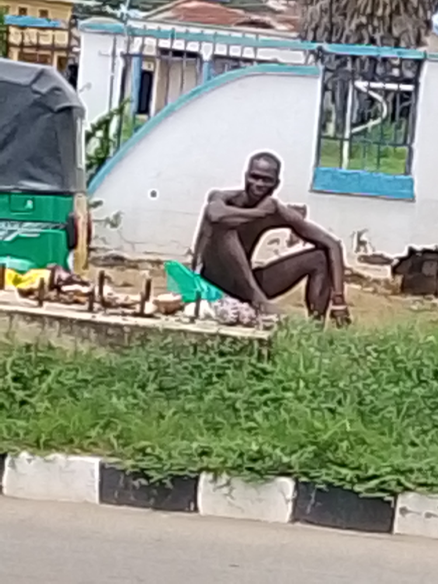 A bare-chested man is sitting on the ground with his legs crossed and arms resting on his knees. He is smiling at the camera. Behind him, there is a green tarp and some other objects, possibly for sale. To the left, part of a green and grey vehicle, possibly a tuk-tuk, is visible. In the background, there is a white building with blue trim and barred windows, and some trees. The scene appears to be outdoors, in what looks like a marketplace or roadside setting. The lighting suggests it is daytime, possibly overcast. The man's expression is one of apparent contentment or amusement. There is no visible text in the image.