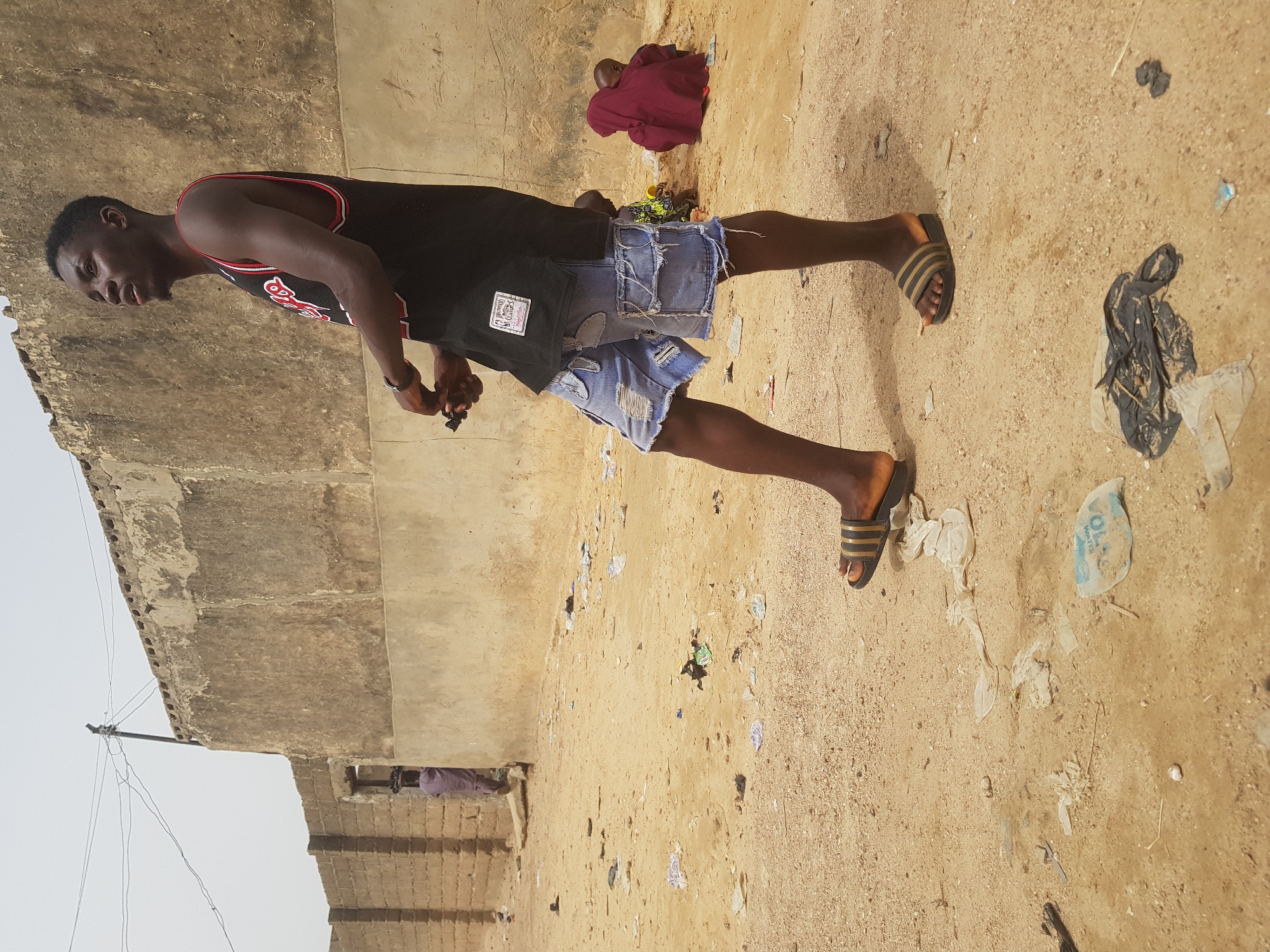 A young man stands in a sandy, littered area near a building in Dutsinma B, Nigeria. He is wearing a black tank top with red trim, torn denim shorts, and striped sandals. He holds a small object in his hands. Debris litters the ground. A figure draped in a red garment is seated on the ground in the background. A building structure is on the left side of the frame.