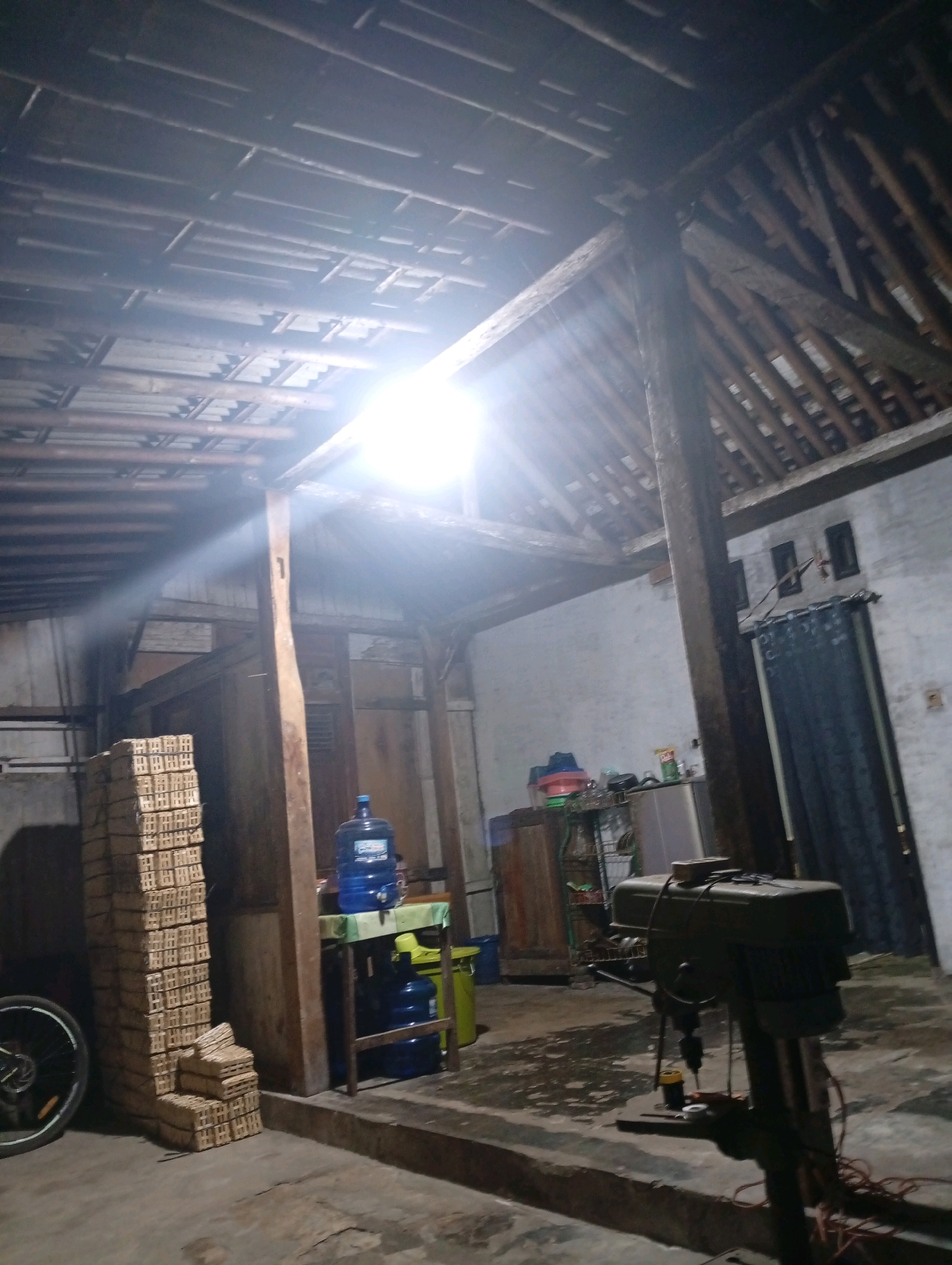 The image depicts the interior of a large, open-air workshop or storage space in Kebanaran, Indonesia. The structure has a high, sloped roof supported by exposed wooden beams and rafters. A bright overhead light source casts strong rays and creates lens flare in the upper portion of the image.
In the foreground to the left, a bicycle wheel is visible. To the right of the wheel, a tall stack of small, rectangular wooden objects, possibly bricks or packaging, stands against a wall. Further to the right, a large blue water cooler bottle rests on a shelf supported by a wooden table. Next to the table, a green trash can is visible.
Dominating the right side of the image is a large, industrial-looking drilling machine. Wires are visible around the base of the machine. In the background, against a white wall, a dark curtain hangs. To the left of the curtain, there are two small, dark rectangular objects mounted on the wall, possibly shelves or electrical outlets. Shelving units with various items, including containers and a refrigerator, are also present in the background. The floor appears to be concrete, with some debris or water present. The overall impression is of a functional, utilitarian space.