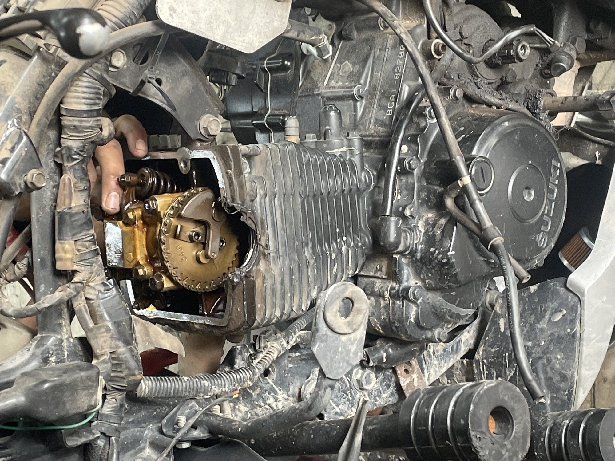 The image shows a partially disassembled motorcycle engine. A hand is visible near the camshaft, which is exposed. Other engine components, including the cylinder head, cylinder block, and crankcase cover, are visible. The environment appears to be a workshop or garage. There is no information to suggest that the image was taken in Dhaka, Bangladesh.