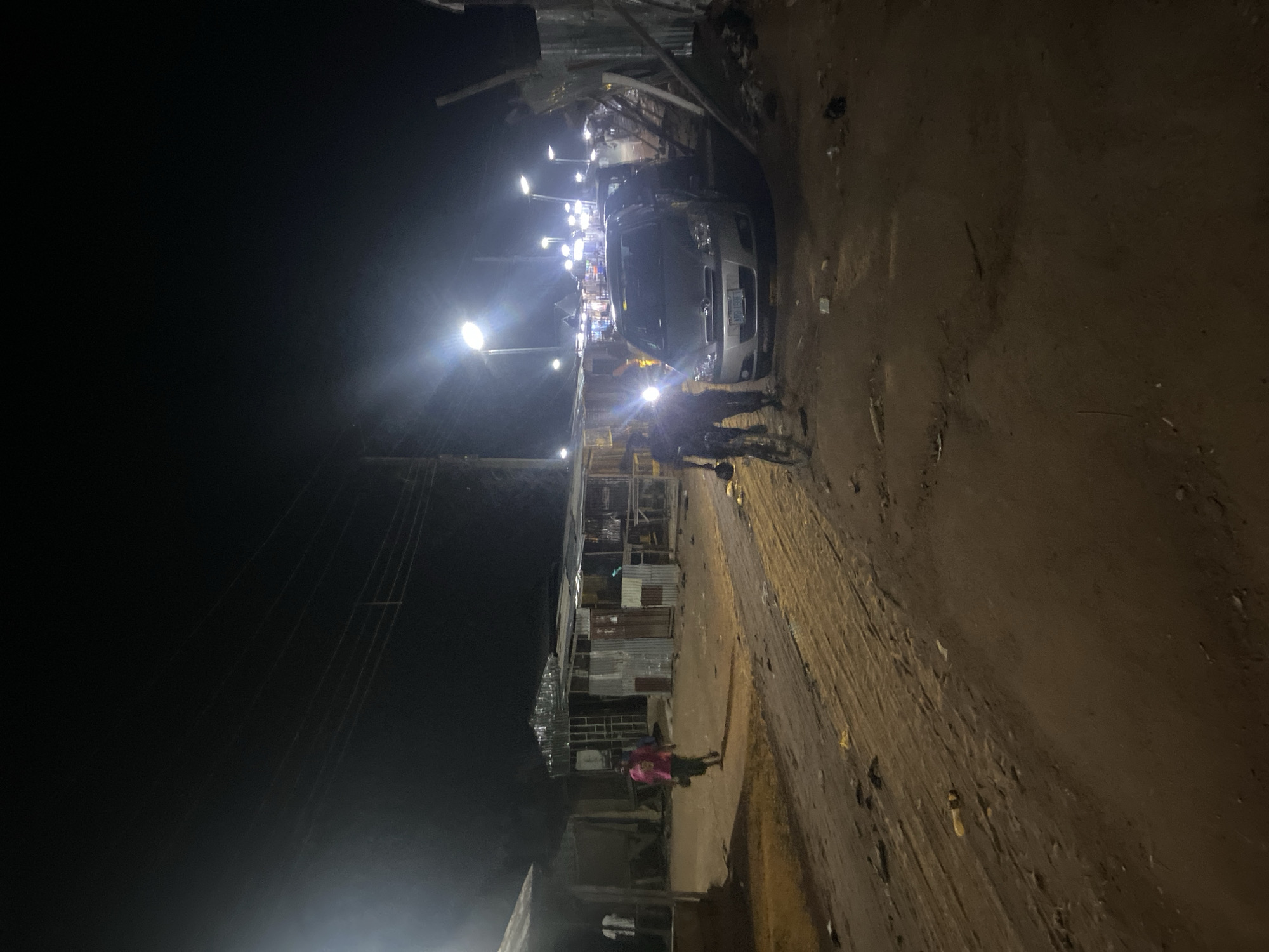 The image depicts a nighttime scene in what appears to be a market or roadside area in Kuson, Nigeria. The primary subjects are two individuals dressed in dark clothing, possibly security personnel, standing near a silver minivan. The minivan is parked on a dirt road, which is uneven and appears somewhat muddy in places.
The setting is illuminated by several bright streetlights, casting harsh light and creating deep shadows. On the left side of the road, there are makeshift structures, likely stalls or small shops, constructed from corrugated metal and other scavenged materials. These structures are arranged in a row, creating a somewhat cluttered and dense environment. The overall impression is of a busy but informal commercial area.
The time of day is clearly night, as indicated by the darkness of the sky and the artificial lighting. There are no clear indications of weather, but the ground suggests it might have rained recently.
A small figure, possibly a child or young person wearing a pink garment, is visible further down the road, adding a touch of human presence to the scene beyond the two individuals near the vehicle. In the distance, more lights suggest further activity or habitation.
There is no visible text in the image that can be deciphered. The overall mood is functional, with the lighting highlighting the utilitarian nature of the structures and the presence of people engaged in what seems to be a routine nighttime activity, possibly security or monitoring. The image captures a glimpse of daily life in a specific Nigerian locality at night.
