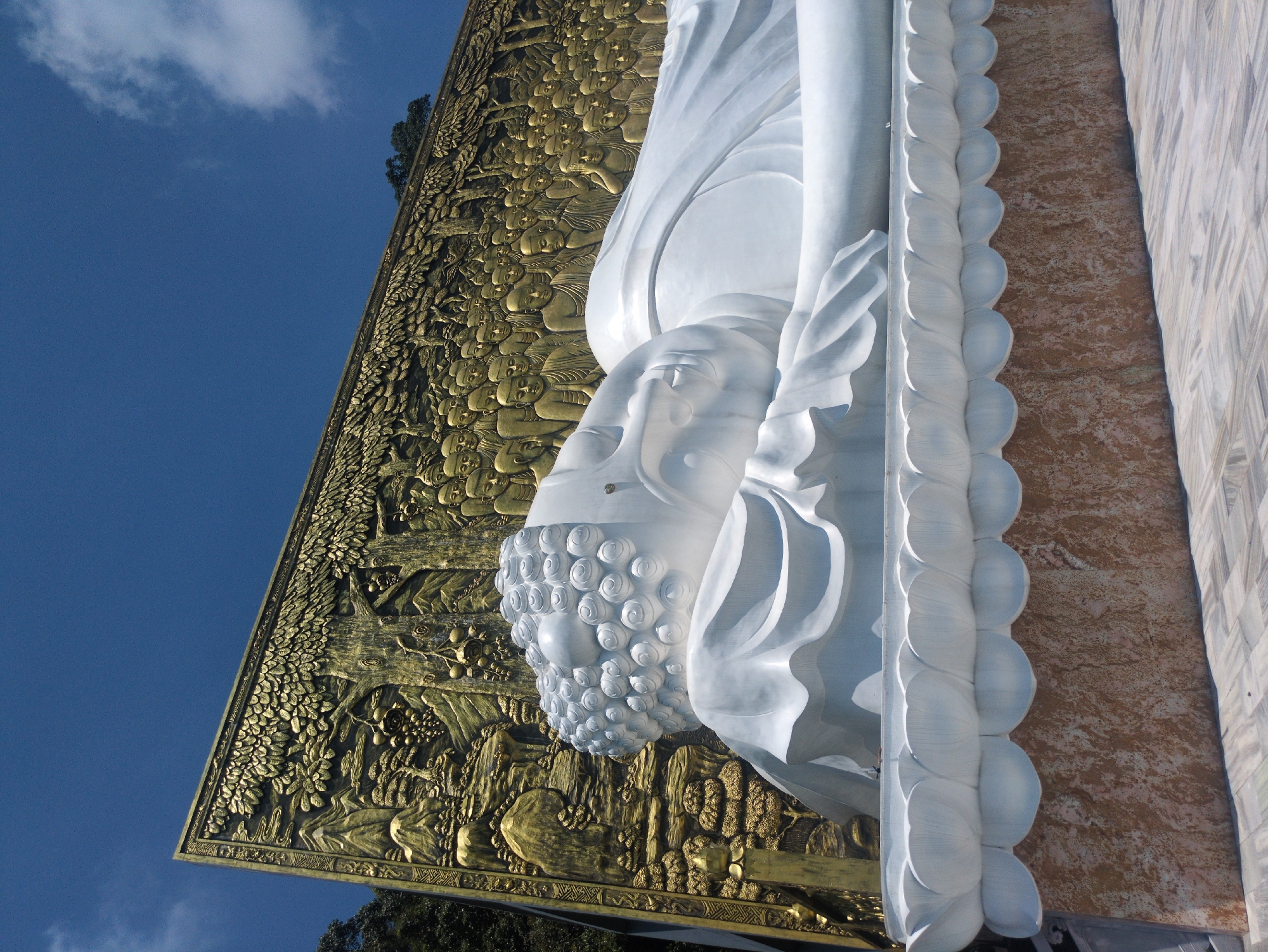 Here is a description of the image.
A vertically oriented, low-angle shot depicts a large, white reclining Buddha statue in Phú Quốc, Vietnam, against a backdrop of a vivid blue sky. The statue's head, shoulder, and draped robes are visible. The Buddha's hair appears as a mass of spiral curls, and the face has a serene expression. Behind the statue is a large golden panel adorned with intricate bas-relief carvings depicting figures and scenes. To the right, behind the statue's body, is a patterned wall of light brown and white shades.