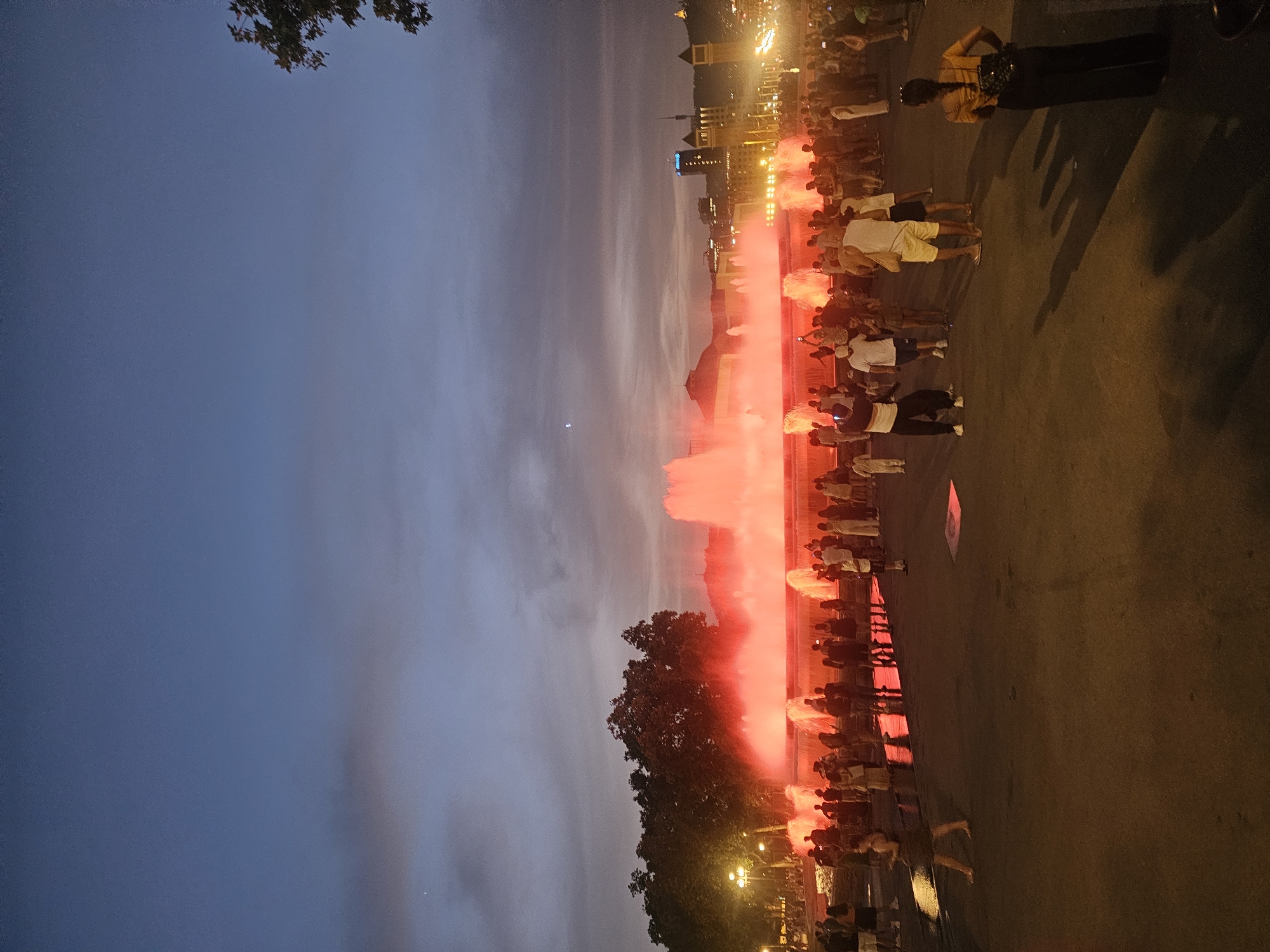 The image depicts a vibrant scene at the Magic Fountain of Montjuïc in Barcelona, Spain, during dusk. The fountain is illuminated with a striking red glow, creating a dramatic visual effect as water jets upwards. A diverse crowd of people, dressed in casual attire, are gathered around the fountain, observing the spectacle. Some are standing, while others are seated, and many are engaged in taking photographs with their mobile phones. The background reveals the city's architecture, with buildings and lights visible in the distance, under a twilight sky with scattered clouds. Trees line the left side of the scene, adding a touch of nature to the urban setting. The ground is paved, reflecting the colorful lights of the fountain. The overall atmosphere is one of public enjoyment and awe at the illuminated fountain display.