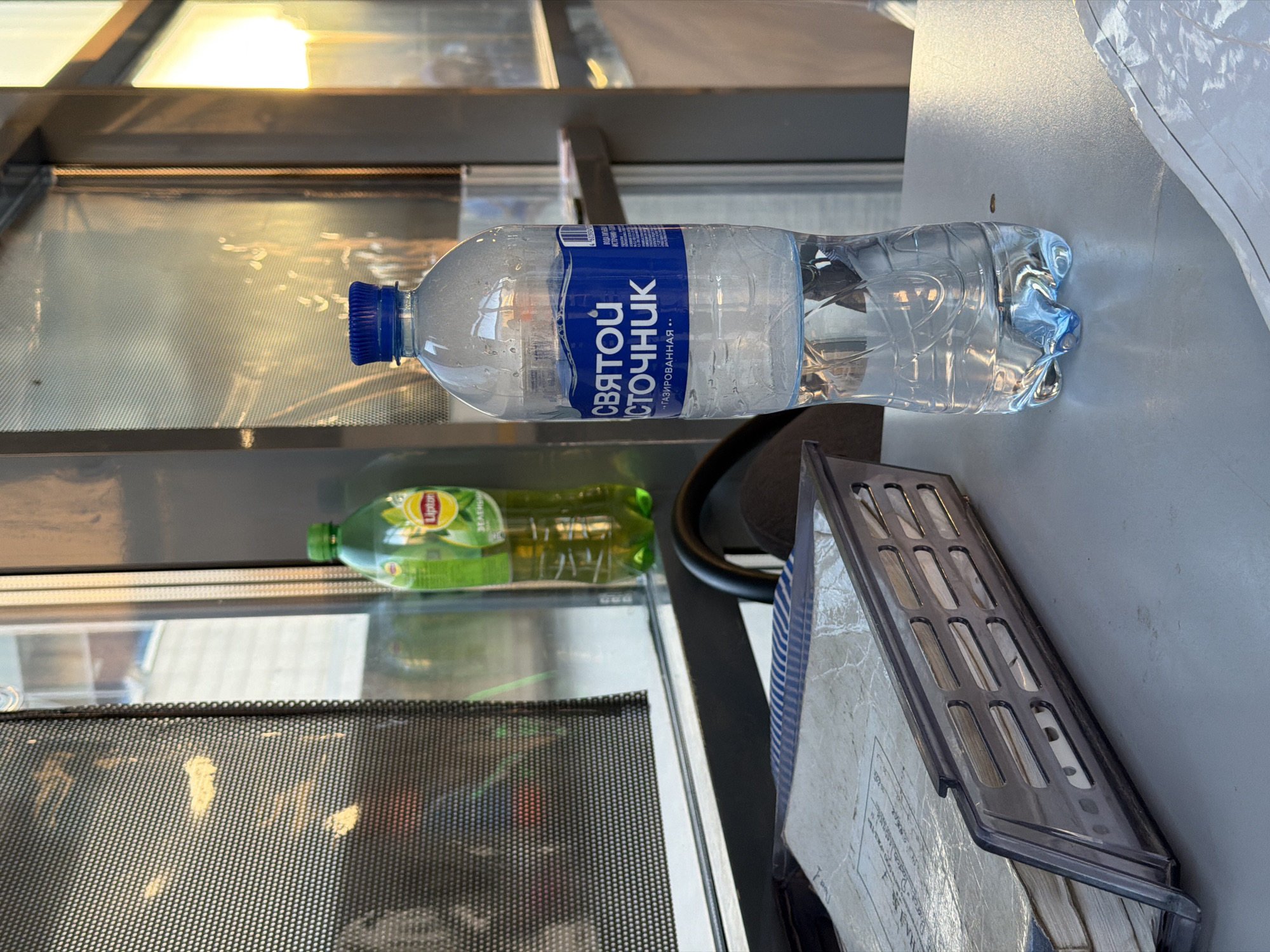A close-up, eye-level shot captures a clear plastic bottle of "Svyatoi Istochnik" mineral water, with a blue label, standing on a light gray counter. The bottle is partially filled with water and condensation. To the left of the water bottle, a green Lipton iced tea bottle is visible, also partially filled. In the foreground, a dark gray, perforated plastic document tray holds several stacks of old, yellowed paper documents. The scene appears to be indoors, with a large window or glass partition in the background that reflects light and shows blurred outdoor elements. The lighting suggests it may be late afternoon or early evening. The overall impression is of a functional workspace, possibly a kiosk or a service counter.
The specific location in Rostov-on-Don, Russia is not identifiable from the visual information provided in the image.