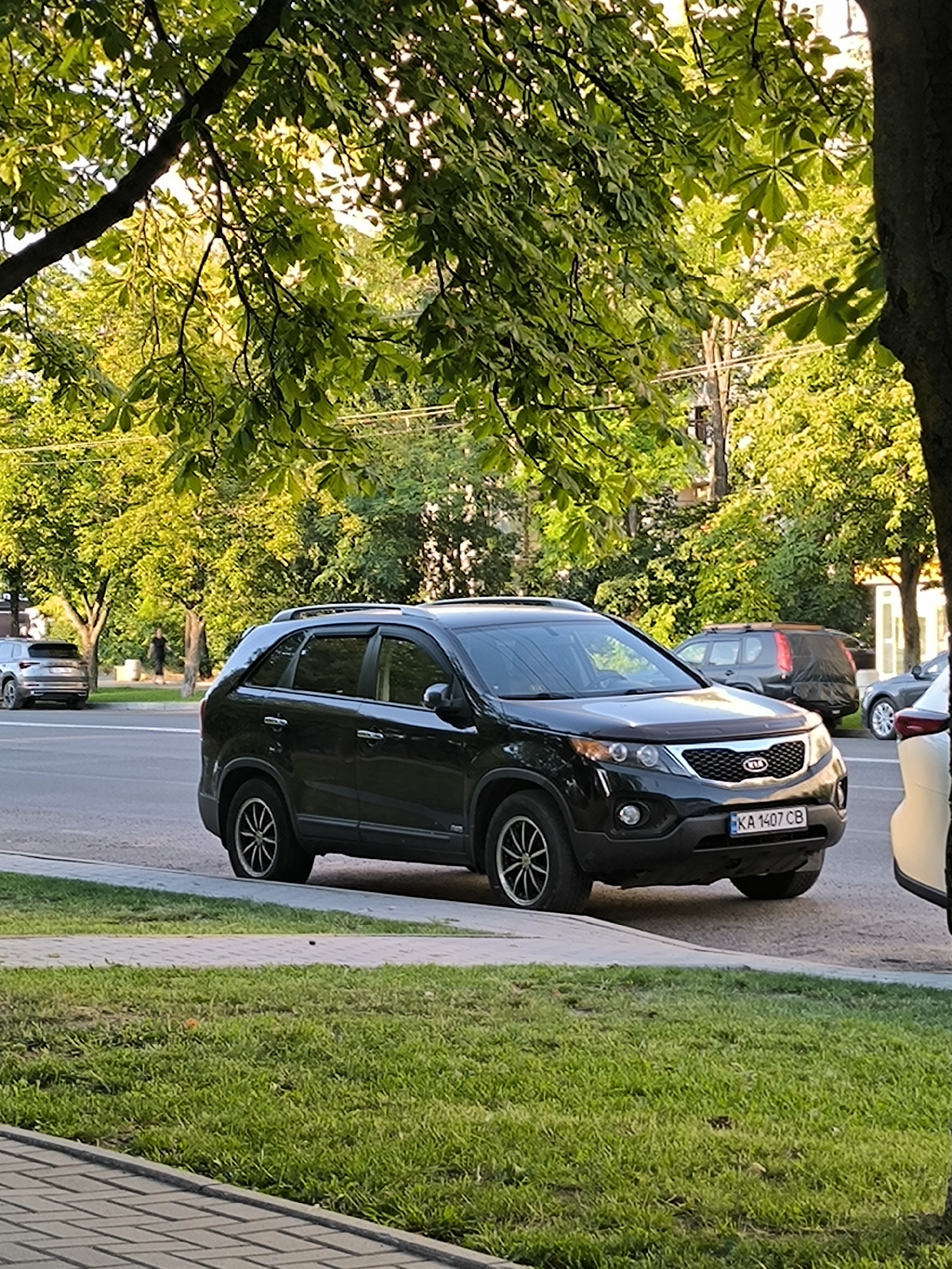A black Kia Sorento is parked on the side of a street in Chisinau, Moldova. The car has a license plate that reads "KA 1407 CB". It is a sunny day, and there are trees with green leaves lining the street. In the background, other cars are visible, as well as a person walking. The foreground shows a grassy area and a paved walkway.