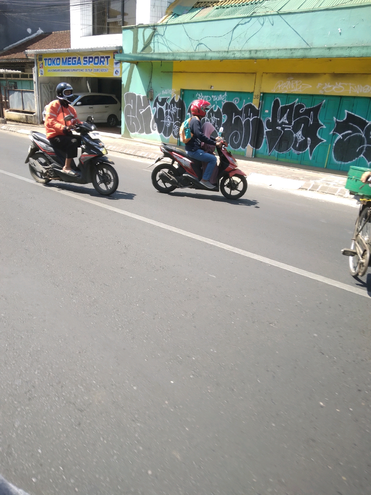A side view of a street in Purwokerto Lor, Indonesia, shows two people riding motorcycles and a person on a bicycle. On the left, a person wearing an orange jacket and a white helmet rides a black motorcycle. On the right, a person wearing a red helmet and a blue shirt rides a red motorcycle. Behind them, a person on a bicycle with a green crate is visible. The background features a building with a yellow and green facade covered in graffiti, and a sign that reads "TOKO MEGA SPORT." The sky is bright and clear, indicating a sunny day.