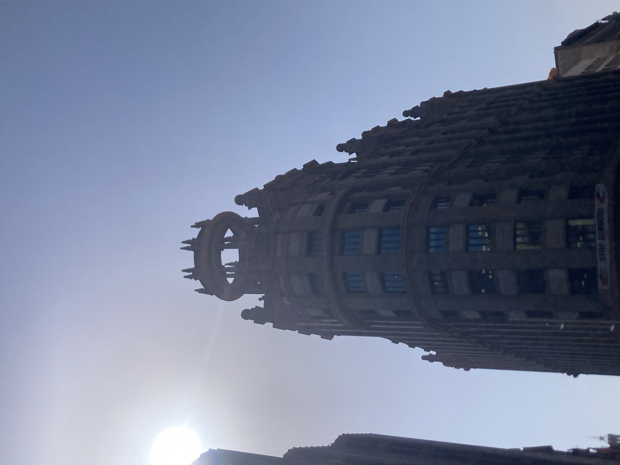 The image, taken from a low angle looking upwards, features the ornate, dark-colored facade of the Casa Amatller building in Barcelona, Spain. The building, a modernist architectural gem designed by Josep Puig i Cadafalch, is characterized by its stepped gable and intricate decorative elements. The sun is positioned in the upper left quadrant of the image, creating a lens flare and casting a bright haze that partially obscures the sky and the top of the building. The sky is a clear, pale blue. To the left, another tall building, likely also in Barcelona, is visible, appearing darker and more uniform in design. The lower portion of the Casa Amatller facade shows windows with dark frames and reflective glass, as well as decorative friezes. A banner with red text is visible at the very bottom of the Casa Amatller, though the text is largely illegible but appears to say "STOP GENOCIDIO". The overall impression is a snapshot of Barcelona's architectural grandeur under a bright, hazy sky.