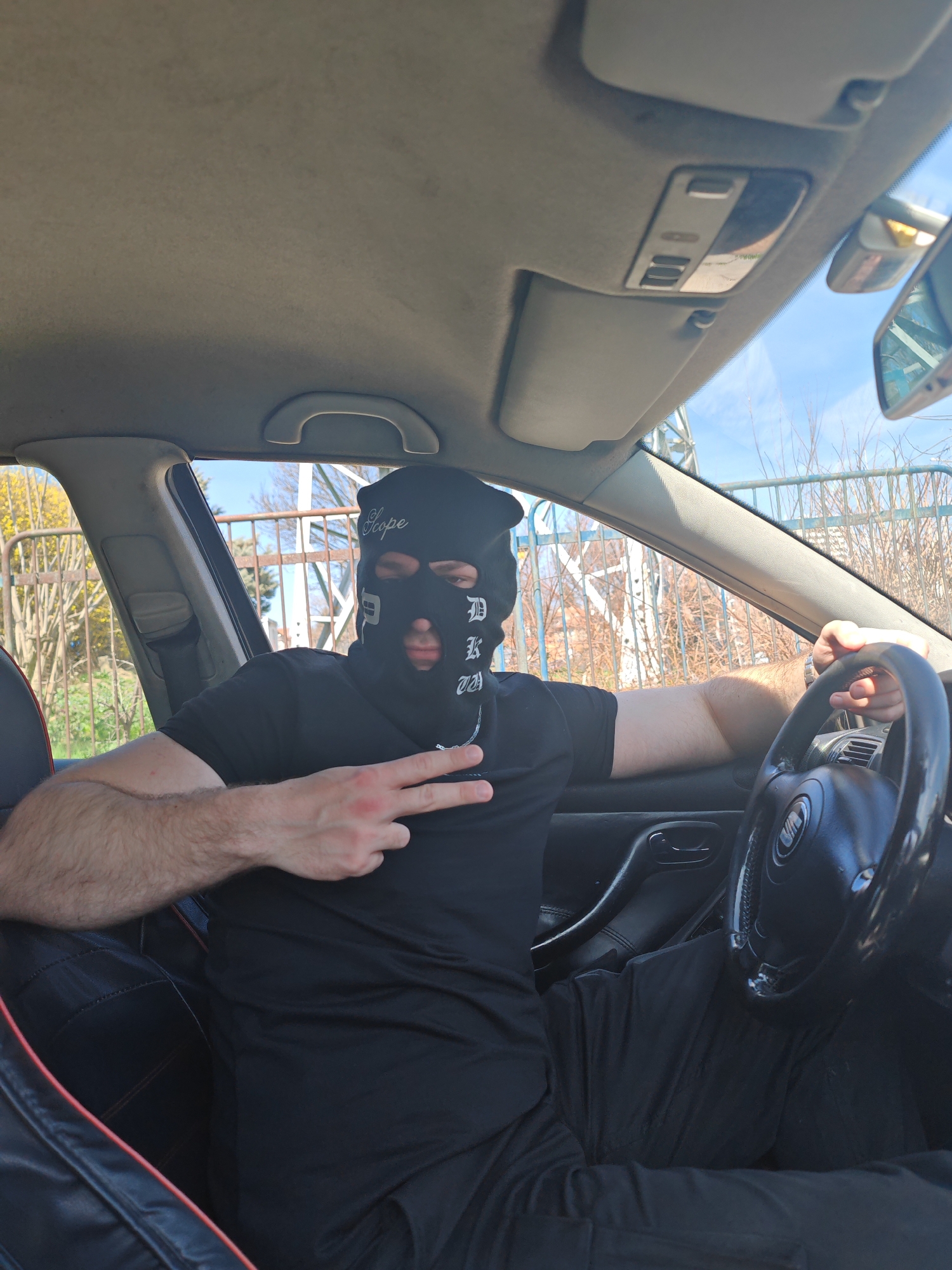 A male individual is depicted in the driver's seat of a black SEAT passenger vehicle, stationary outdoors in Shumen, Bulgaria. The individual wears a black balaclava covering the entire head, featuring "Scope" printed on the forehead and stylized "DKW" on the right cheek. They are dressed in a black short-sleeved t-shirt and black pants. The left hand, positioned near the chest, forms a two-finger peace sign. The right hand rests on the black steering wheel, displaying a watch on the wrist. The vehicle's interior includes a light gray headliner, an overhead console, and black seats with red piping visible on the driver's seat. Through the windows, a light-colored metal fence, leafless deciduous trees, and yellow flowering bushes are visible under a clear blue sky, indicating daylight conditions.