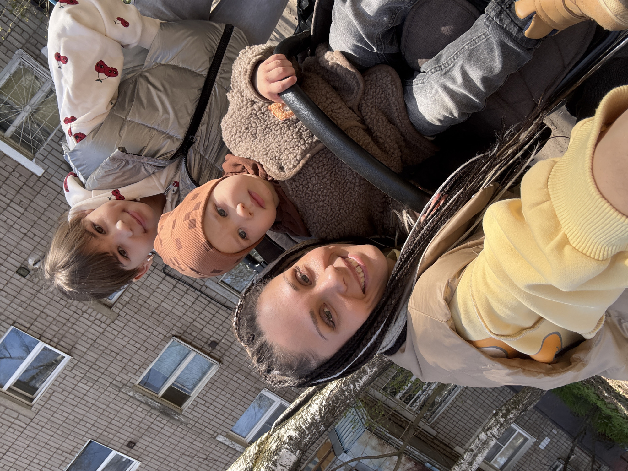 A woman with braided hair takes a selfie with two children outside of a brick building. The older child is wearing a white hoodie with cherries on it and a silver jacket. The baby is wearing a brown hat and jacket and is sitting in a stroller. The building has windows with white frames. The woman is wearing a yellow sweater and a tan jacket. The image was captured in Rusanovo, Russia.