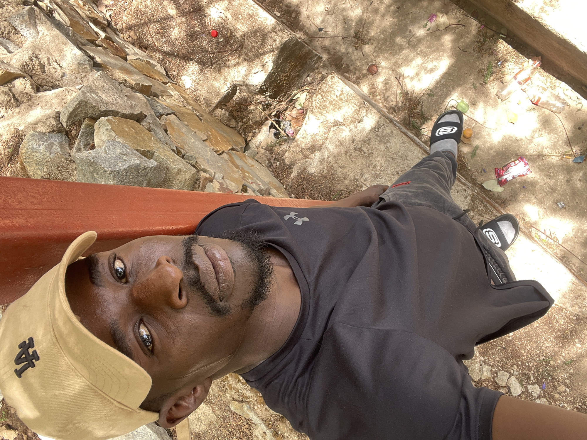 A male subject takes a selfie outdoors, likely in Jos, Nigeria, looking directly into the camera from a low, overhead angle. He has dark skin and wears a tan baseball cap with an "LA" logo, a black short-sleeved t-shirt bearing an Under Armour logo, dark grey sweatpants, and black Skechers slide sandals over grey socks.
The background presents an unkempt outdoor scene. To the left, a natural rock formation with various sizes of light-colored rocks is visible. To the right, the ground transitions to concrete ledges or steps, interspersed with loose dirt, dried leaves, and debris. Scattered across the ground are several pieces of litter, including clear plastic bottles, food wrappers, a red bottle cap, and what appear to be small fallen fruits.
A prominent orange or reddish-brown structure, possibly a wooden railing or bench, runs across the lower portion of the image, close to the subject. The lighting indicates a bright, sunny day with strong, distinct shadows, suggesting midday or early afternoon. The overall setting combines natural elements with neglected man-made structures and litter.