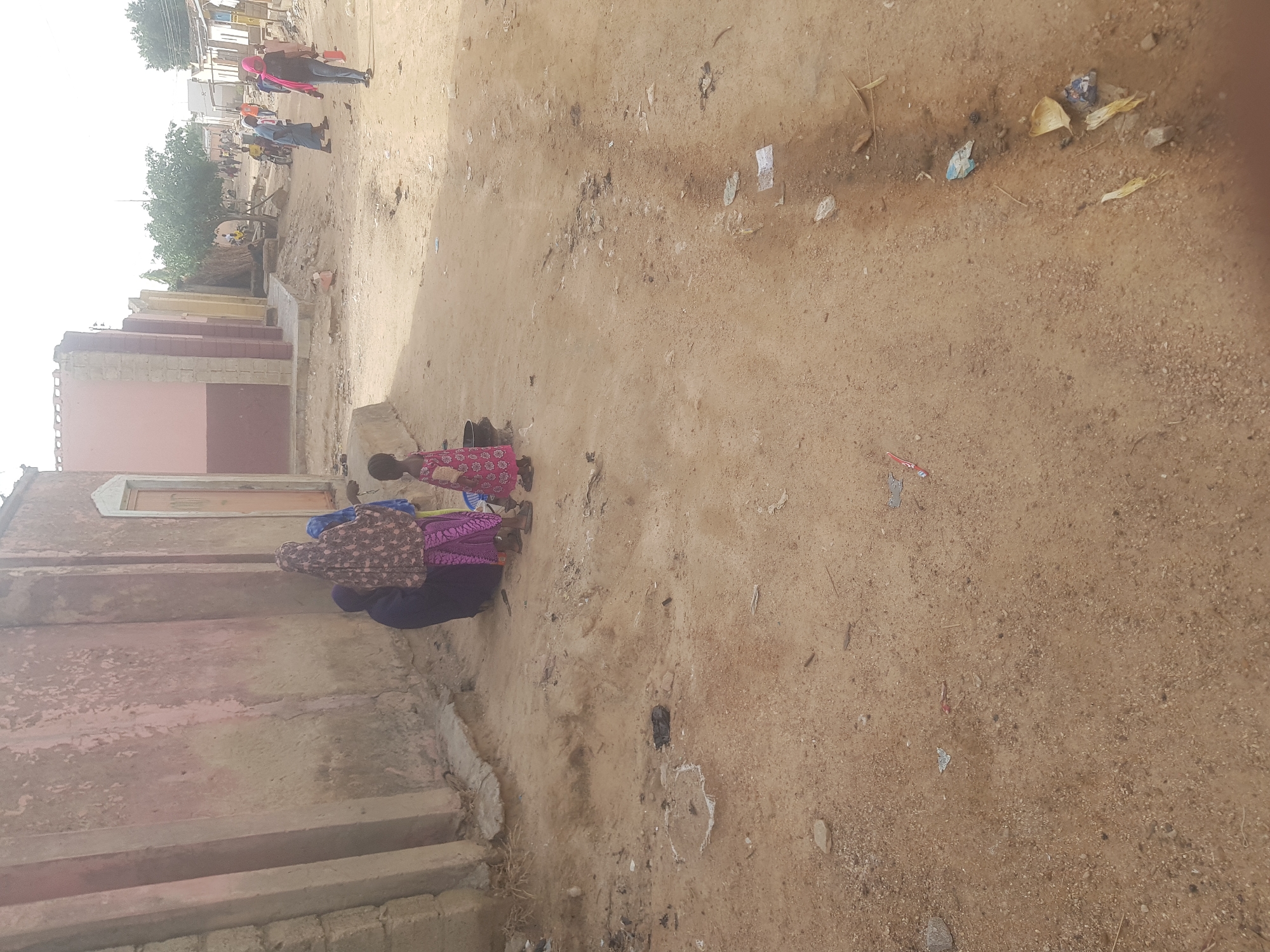 The image captures a scene in Dabawa, Nigeria, showing an unpaved street alongside a building. The ground is sandy and scattered with debris. Adjacent to the building, a group of people are present, including a child in a red and pink patterned dress. The adult figures are clothed in traditional attire, one with a brown patterned fabric and another in purple. In the background, further down the street, more individuals are visible, some wearing brightly colored garments. A line of buildings and trees are present in the far distance, adding depth to the urban environment.