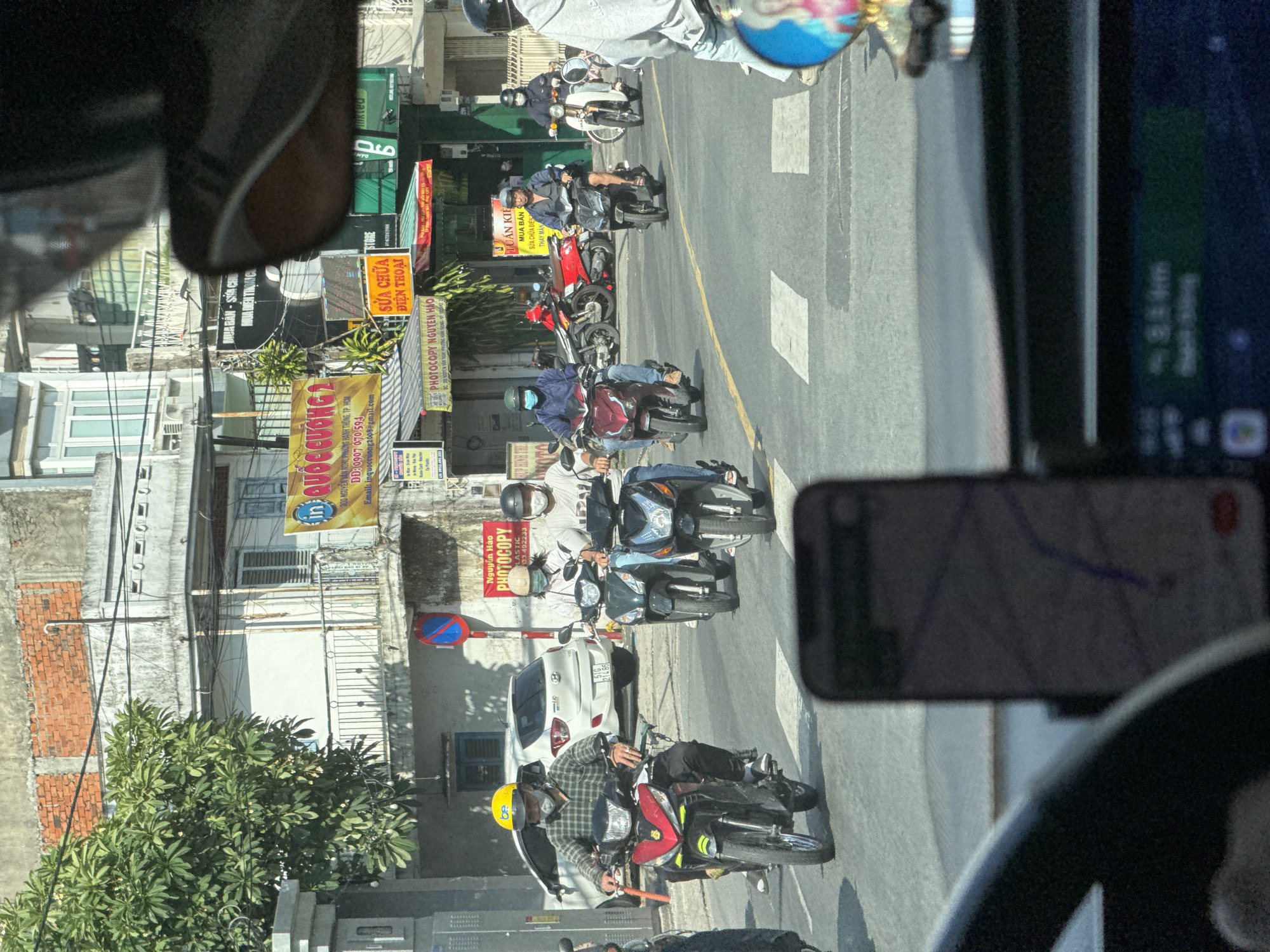 The image captures a street scene in Thủ Đức, Vietnam, likely during daylight hours. The perspective is from inside a vehicle, looking forward. Several motorbikes are prominent on the road, many with riders wearing helmets and face masks. One motorbike in the foreground, a red and black model, is ridden by a person wearing a yellow helmet and a plaid shirt. To the right, two other motorbikes are visible, one dark green and one black, with riders also masked. Further down the street, more motorbikes and a white car are present. The surrounding environment includes buildings of varying architectural styles, some with signs in Vietnamese. One prominent sign reads "QUỐC CƯỜNG 2" and another advertises "COPY Nguyen Hao". The road is paved and marked with pedestrian crossings. A smartphone mounted on the dashboard displays a navigation interface.