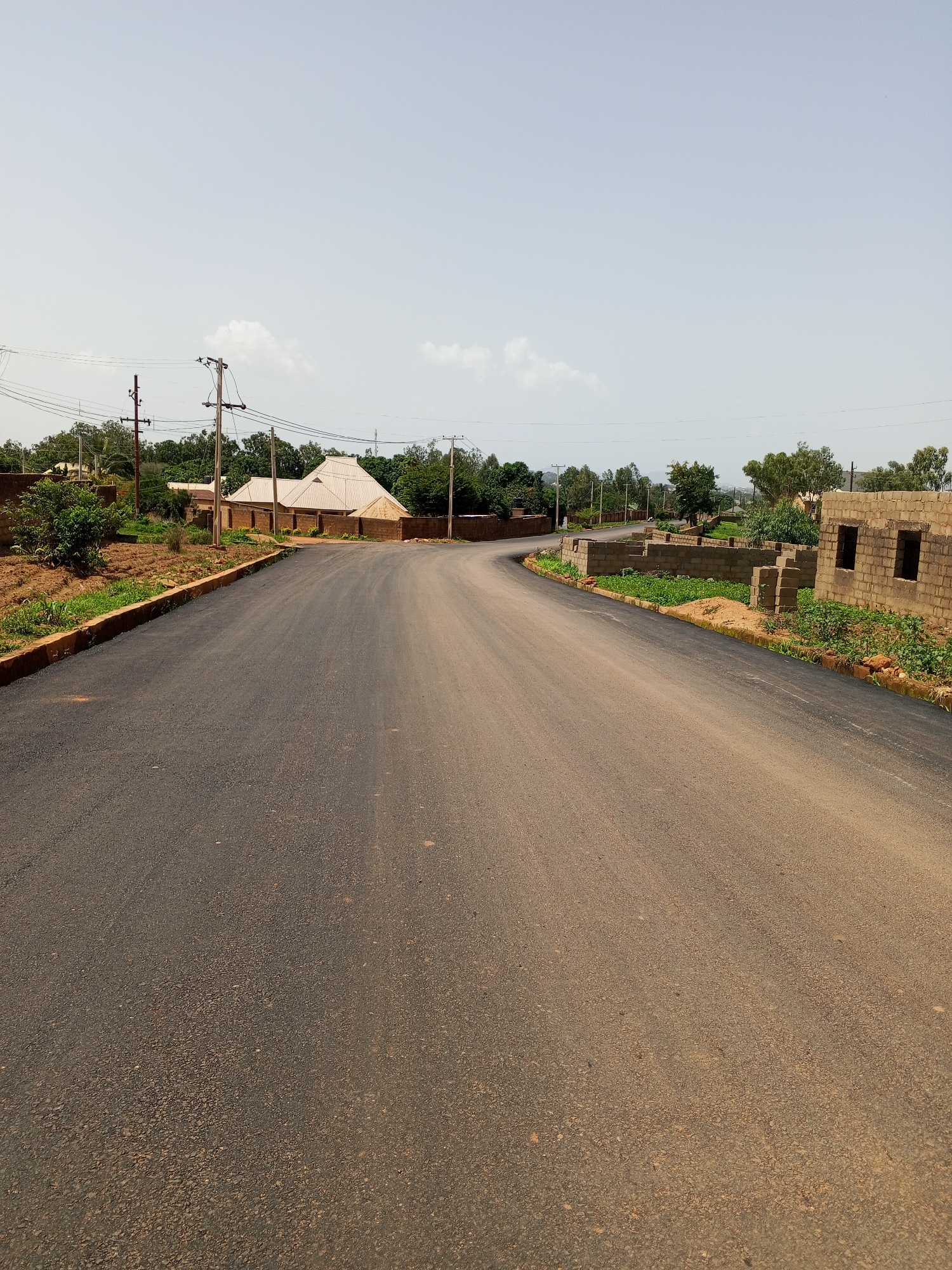 The image depicts a paved road curving through a semi-urban or rural landscape. The road surface is asphalt, showing signs of recent construction with fresh asphalt along the edges. The scene is set in Bukuru, Nigeria. The time of day appears to be late morning or early afternoon, indicated by the bright, clear sky with a few scattered clouds. The weather is fair and sunny.
Along the left side of the road, there are areas of cleared land, some with sparse vegetation, and a small bush. Further back, a building with a distinctive, multi-tiered metallic roof is visible, surrounded by a brick wall. Utility poles with electrical wires are present along the roadside, suggesting access to electricity.
On the right side of the road, construction of brick buildings is underway, with walls built from concrete blocks but lacking roofs and windows. Green vegetation is growing around the construction sites. In the background, a line of trees and foliage stretches across the horizon, with more buildings and utility poles visible further in the distance. The overall impression is one of development and ongoing construction within a natural setting. No people or specific activities are discernible in the image. There is no visible text.