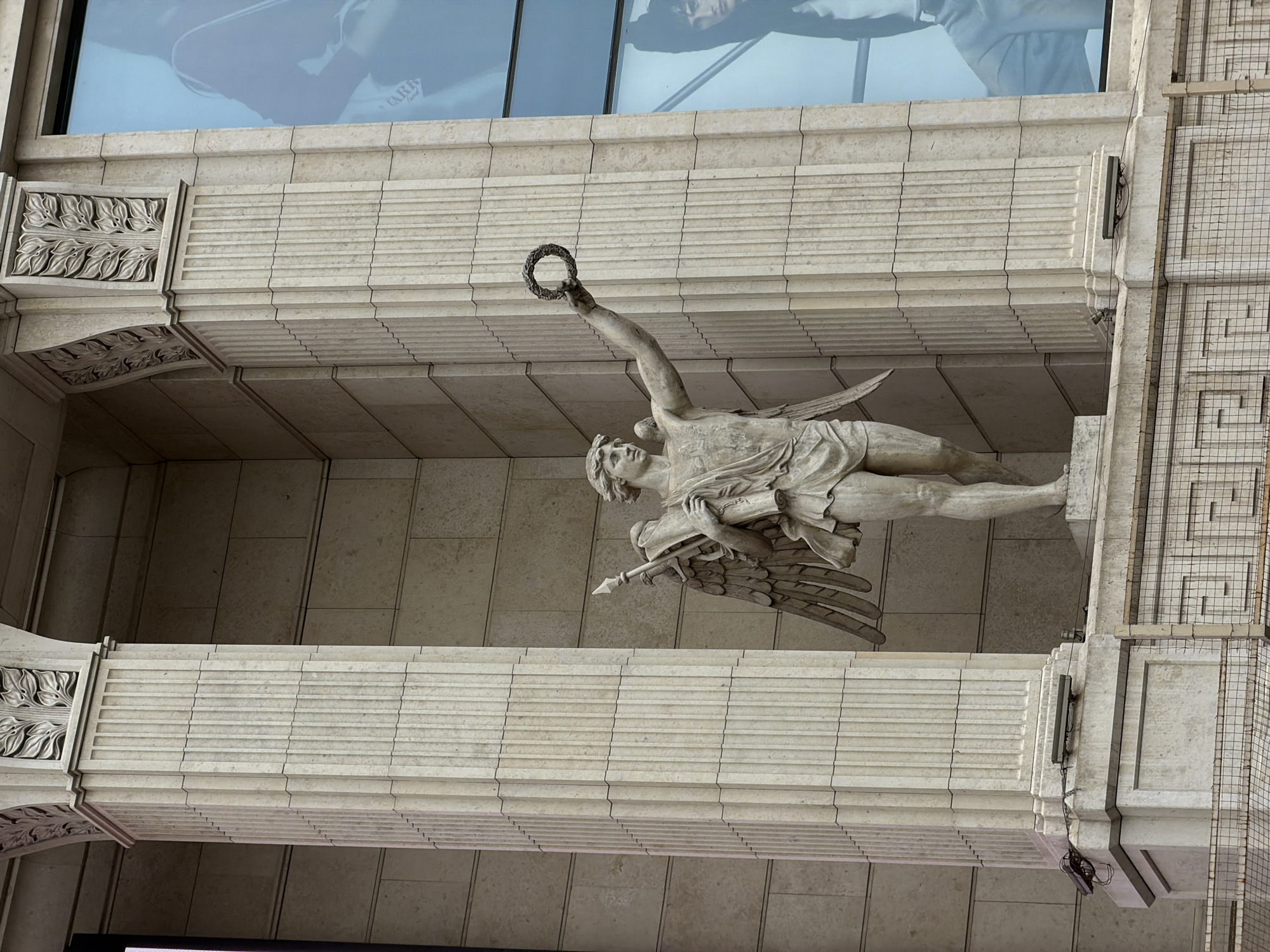 The image depicts a statue of a winged figure, likely an angel or allegorical figure, mounted on a building in Saint Petersburg, Russia. The statue is made of light-colored stone and is depicted horizontally, suspended between two architectural elements of the building's facade. The figure is nude except for a drape across the lower body. The figure's right arm is raised, holding a wreath aloft. The statue holds a large spear in its left arm. The building has a classically inspired design with detailed carved ornamentation. There is a reflection of modern buildings in windows above. A wire grid is also visible on a portion of the building.