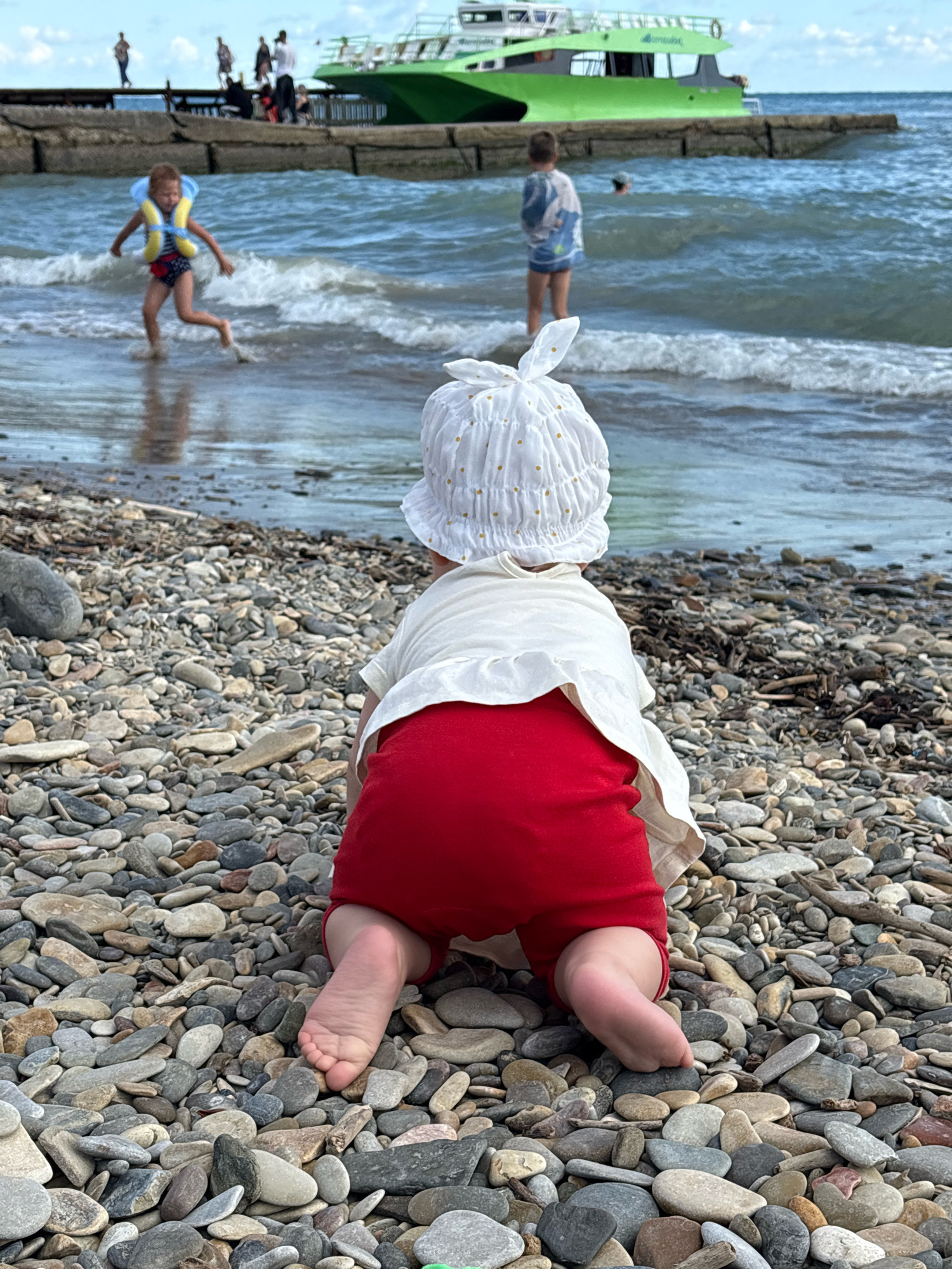 The image depicts a pebbled beach in Arkhipo-Osipovka, Russia, with the Black Sea in the background. In the foreground, a baby wearing red shorts and a white hat is crawling on the pebbles. In the middle ground, a young child in a life jacket is playing in the shallow water near the shore, with another child standing further back, facing away from the camera. In the background, people are visible on a pier, and a green passenger boat is docked. The sky is partly cloudy.