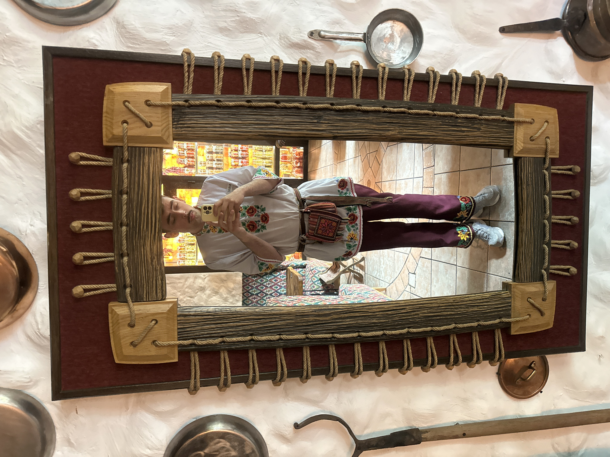 The image shows a full-length mirror with a decorative, rustic frame made of wood and rope. A young person with a mustache is reflected in the mirror, taking a selfie with a smartphone. They are wearing a traditional-style white shirt with embroidered floral patterns around the collar and hem, burgundy trousers also with embroidered details at the ankles, a woven belt with a small pouch attached, and grey sneakers. The background reflected in the mirror shows an interior with patterned seating and shelves stocked with jars, suggesting a restaurant or cultural establishment. The wall on which the mirror hangs is textured and white, with various metal kitchen utensils and decorative items mounted on it, including pans and a long-handled tool. The overall style suggests a Ukrainian cultural theme. The specific location in Ternopil, Ukraine cannot be determined from the image alone.