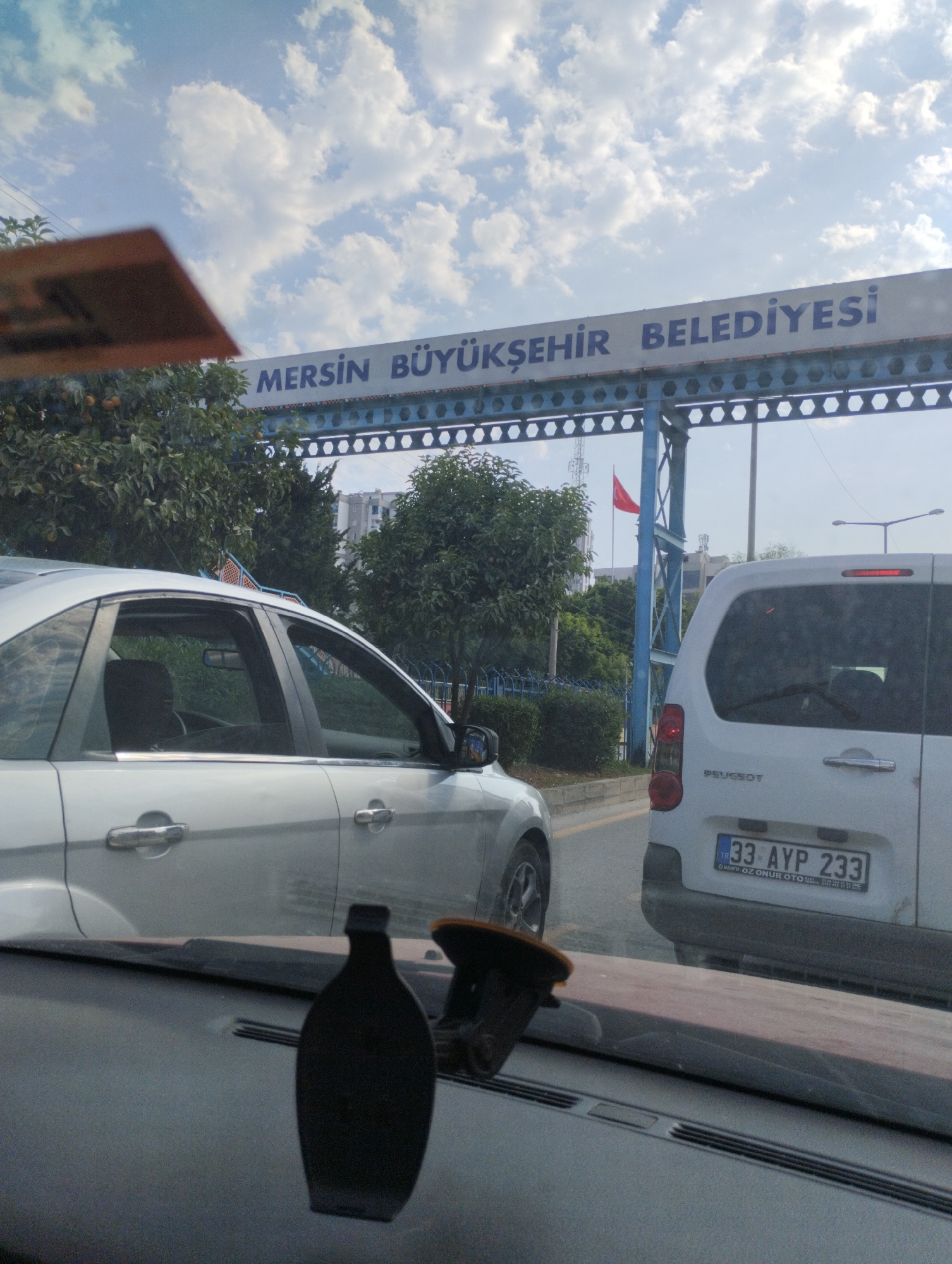 The image shows a view from inside a car, looking forward through the windshield. In the foreground, the dashboard and part of the steering wheel are visible, along with a black decorative item. Ahead, there are two cars in traffic. The car on the left is a silver sedan, and the car on the right is a white van. The white van has a license plate that reads "33 AYP 233" and "TR". Above the cars, a blue archway with the text "MERSİN BÜYÜKŞEHİR BELEDİYESİ" is visible. Behind the archway, a Turkish flag is flying from a flagpole. Trees and buildings are visible in the background. The sky is partly cloudy. This appears to be a scene at an entrance or checkpoint for the city of Mersin, Turkey, likely on a road in Mezitli.