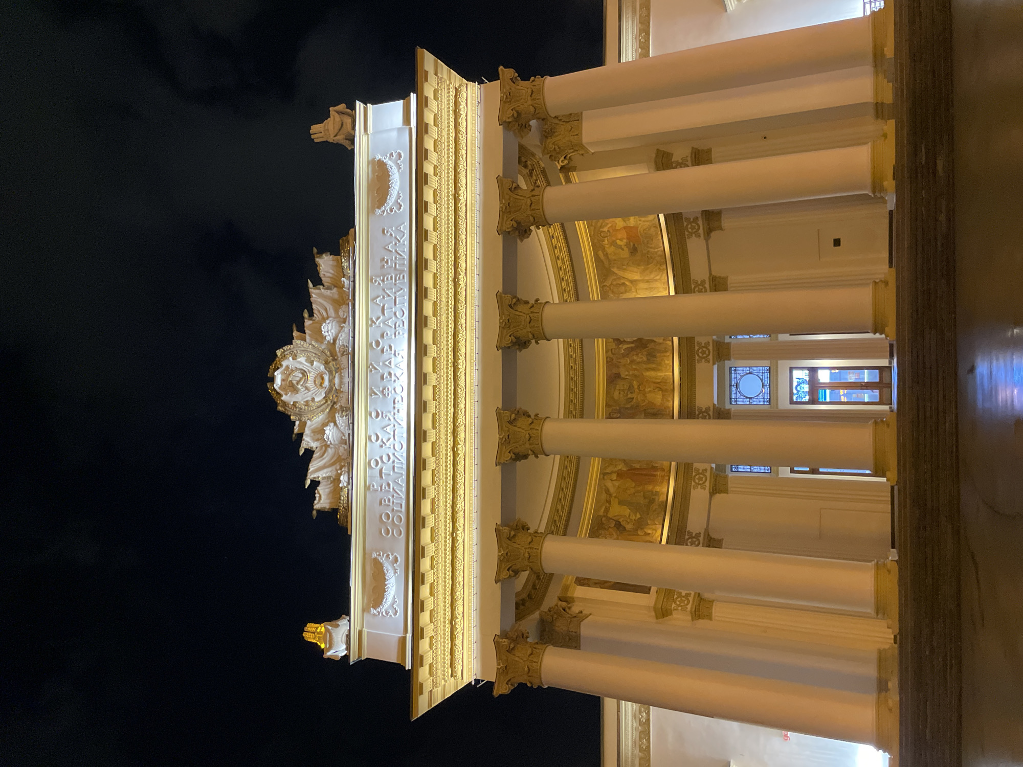 This image depicts a building in Moscow, Russia, specifically the Central Pavilion at VDNKh (Exhibition of Achievements of National Economy). The photograph, taken at night, shows a portion of the building's illuminated facade against a dark sky.
Key features include:
* **Architectural Style:** The building features neoclassical architecture with prominent columns and ornate decorative elements.
* **Columns:** There are multiple white columns with decorated Corinthian capitals.
* **Facade Detail:** The facade is intricately adorned with Soviet-era symbols and inscriptions, including the Order of Lenin and the words "Р. СОВЕТСКАЯ СОЦИАЛИСТИЧЕСКАЯ" (Russian Soviet Socialist).
* **Lighting:** The building is brightly lit, highlighting its architectural details.
* **Windows:** Windows can be seen in the background between the columns.
* **Murals:** Murals can be seen on the walls behind the columns.