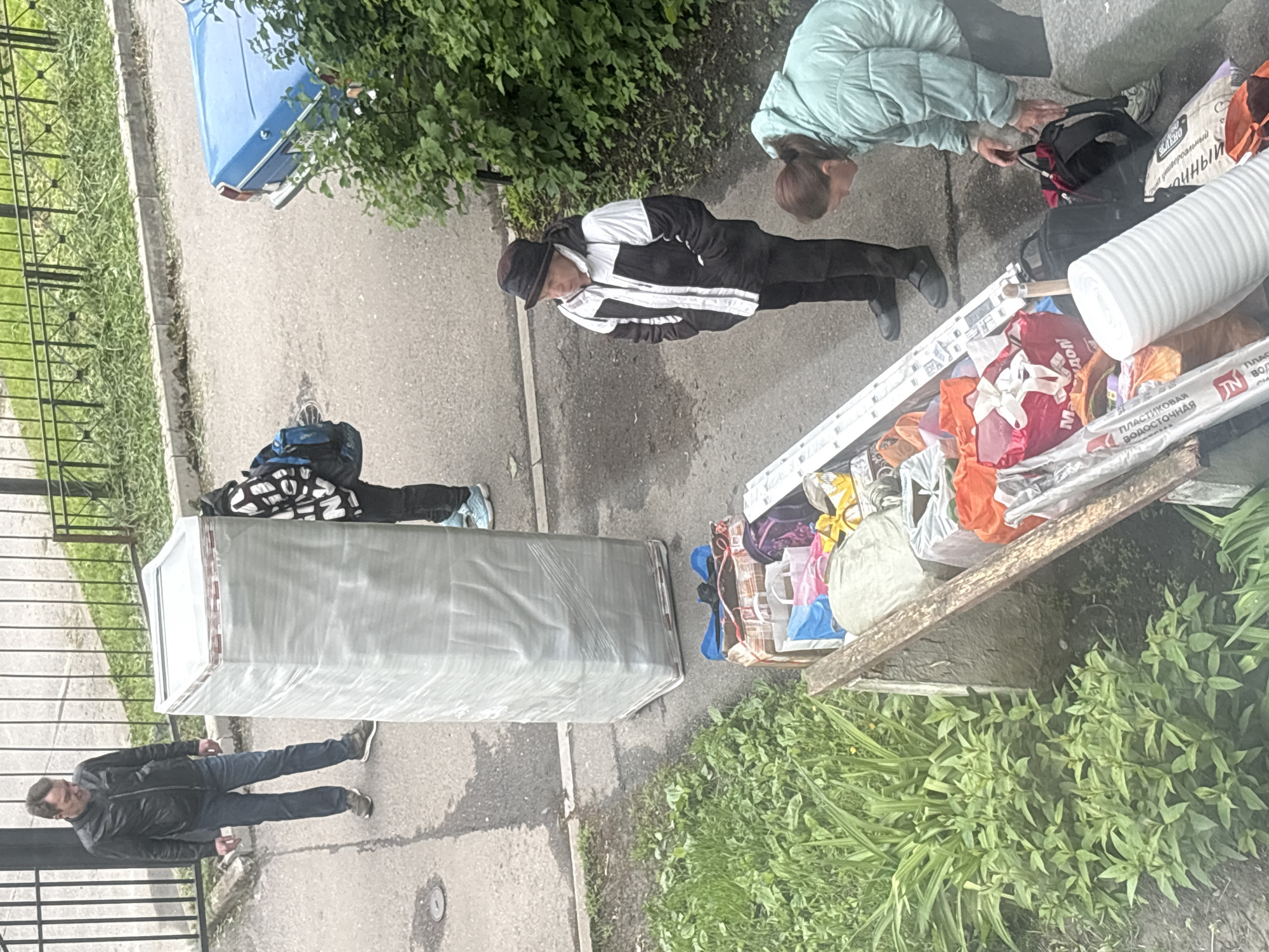 A street scene in Saint Petersburg, Russia. Four people stand on the sidewalk near a curb. To the left of the image is a large rectangular item wrapped in gray packaging film. Next to it is a mound of possessions spilling from an open wooden box. Near the possessions, a woman in a light green jacket kneels and appears to be adjusting a black backpack. A man in a black and white jacket and cap stands near her, looking down at the possessions. To the left of the rectangular package, a person in a patterned jacket and blue jeans stands, and further left is a man in a black jacket and blue jeans. A blue car is parked in the background, obscured by greenery. A metal fence is visible on the far left of the frame. The setting appears to be a residential area with a mix of paved surfaces and vegetation.