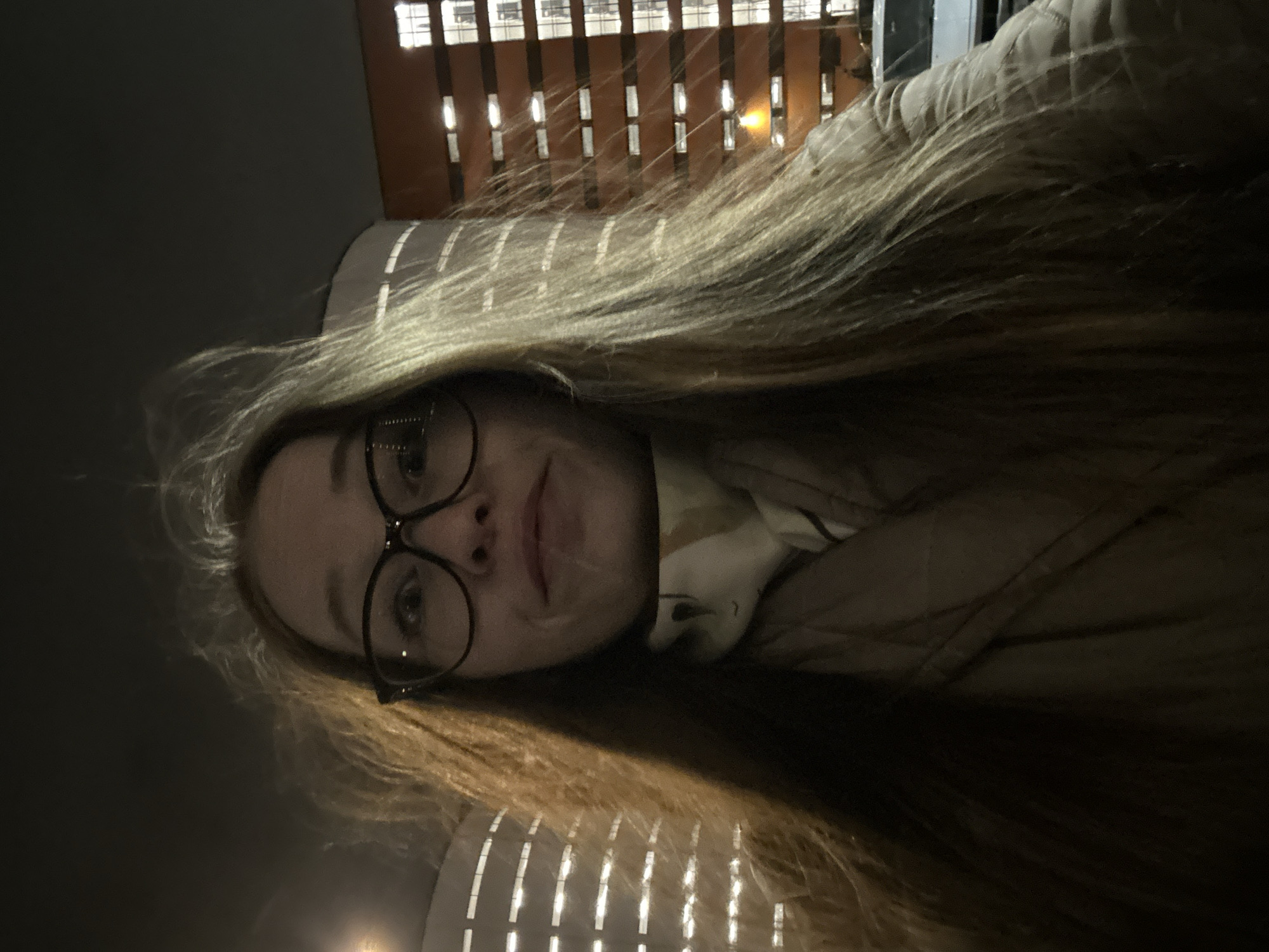 A close-up photo, rotated 90 degrees clockwise, features a woman with long, light brown hair and dark-framed round glasses. Her face is directed towards the camera with a neutral expression, possibly a slight smile. She appears to be wearing a light-colored jacket or coat, possibly quilted. The setting is outdoors at night in an urban environment, likely in Saint Petersburg, Russia. In the background, there is a building with numerous illuminated windows, casting light and creating a grid-like pattern of vertical and horizontal lines. The overall lighting is dim, with the main light source appearing to be from the front, possibly a phone screen or flash, along with ambient light from the distant building. No visible text is present in the image.