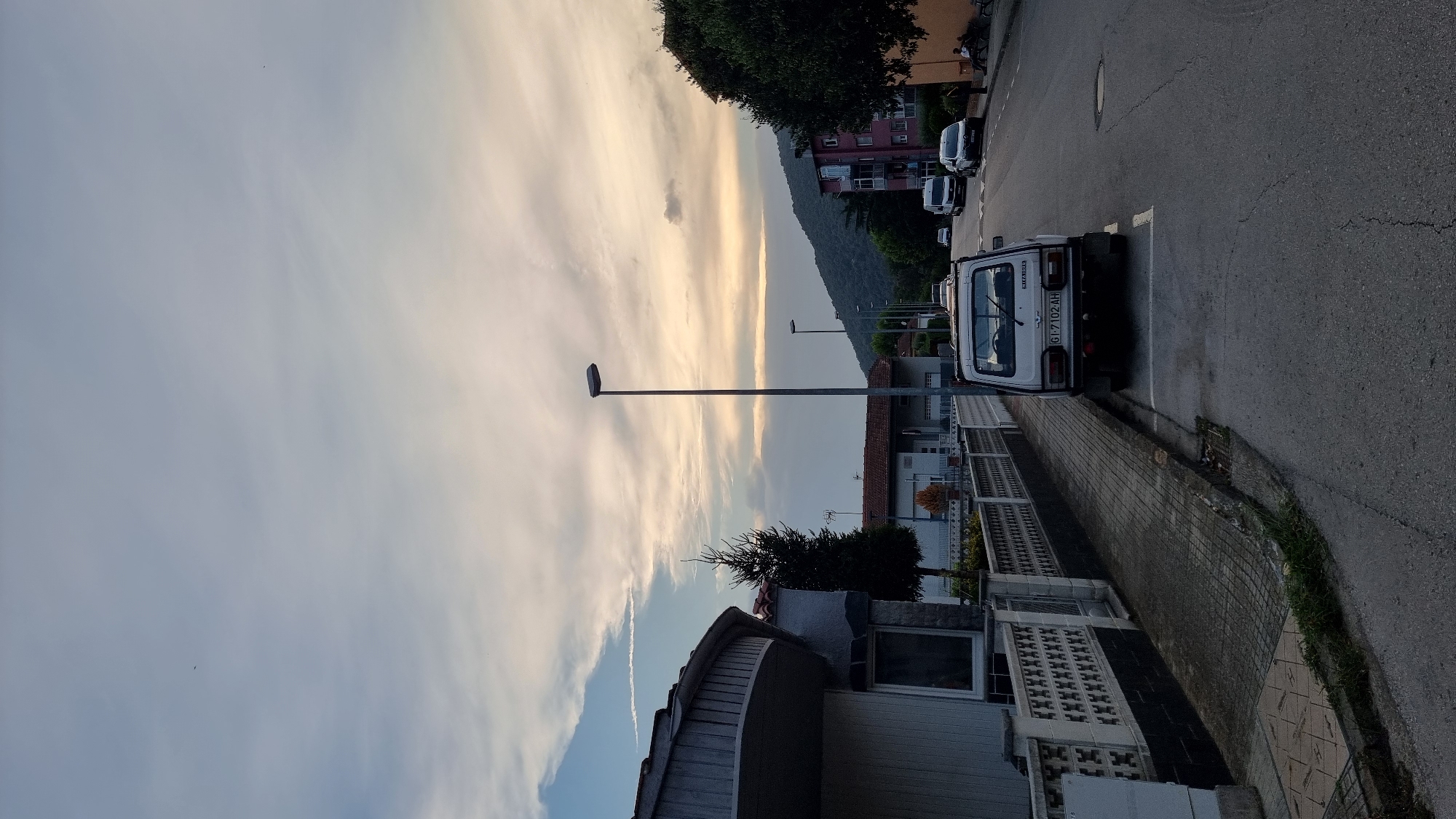 The image depicts a street scene in Sant Joan Les Fonts, Spain. The sky is filled with clouds, transitioning from a light blue at the top to a pale yellow near the horizon, indicating either dawn or dusk. A single contrail is visible in the upper left portion of the sky.
In the foreground, a paved road runs from the bottom right corner towards the upper left. To the left of the road is a sidewalk with a patterned tile surface, bordered by a concrete wall with a decorative lattice pattern. A white car, possibly a Lada Niva, is parked on the right side of the road, facing away from the viewer. Its license plate reads "GI-7102-AH". Further down the road, other vehicles are visible, including white vans and a darker car.
On the left side of the street, a modern-looking house with a curved roofline and white siding is partially visible. Adjacent to it, a tall, slender evergreen tree stands next to a low-rise building with a tiled roof. Beyond these, more residential buildings with tiled roofs line the street. To the far right, a hillside covered in dense green trees rises against the sky, with a few buildings nestled among them. A street lamp stands prominently on the right side of the road, and another is visible further down. A few people are seated on a bench on the right sidewalk in the distance. The overall impression is a quiet residential street during a transitional time of day.