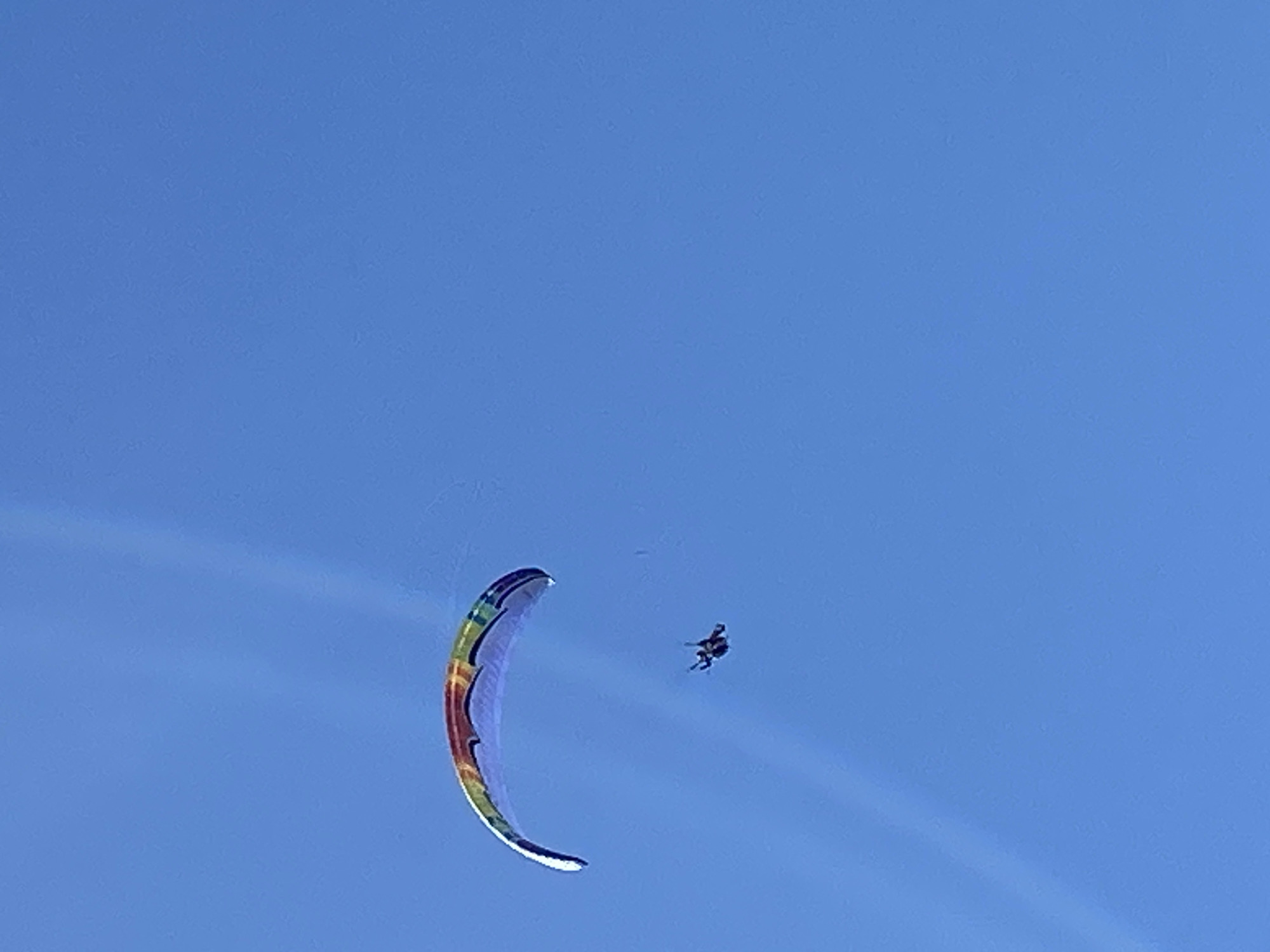 This image captures a paraglider high in a clear blue sky. The sole subject is a person engaged in paragliding. They are seen suspended below a large, colorful paraglider wing, which is the main object in focus besides the person.
The paraglider wing is distinctively vibrant, featuring a rainbow-like spectrum of colors including red, orange, yellow, green, and blue segments, arranged in a striped pattern along its curved surface. It has the characteristic crescent or arc shape of a ram-air canopy, suggesting it's designed for aerial sport. The person appears as a small dark silhouette beneath the expansive wing, indicating they are at a considerable altitude.
The scene is set against a backdrop of an uninterrupted, bright blue sky, with no clouds visible. This suggests a clear, sunny day, ideal for paragliding. The image conveys the activity of aerial flight, with the person actively engaged in controlling the paraglider. There are no other visible objects or features besides the paraglider and the individual.
No specific time of day can be determined beyond it being daylight, and no emotions are discernible from the distant view of the person. There are no visible textual elements in the image. The provided context indicates the location is Boreti, Montenegro, implying this aerial activity is taking place above this area.
